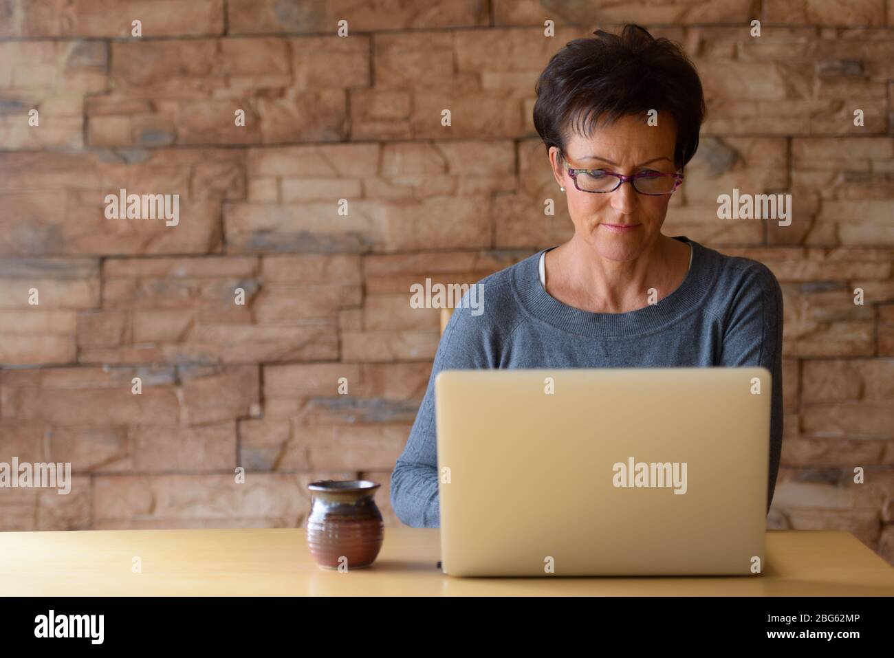 Reife schöne Frau trägt Brillen und Laptop auf Holztisch gegen Ziegelwand Stockfoto