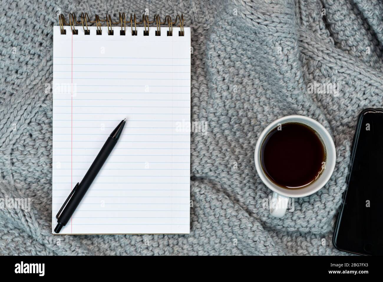 Flaches, gemütliches Schreibhaus mit warmem Pullover, Kaffee, Notizpapier und Smartphone. Arbeiten Sie von zu Hause aus. Schreiben. Warmes Konzept bei kaltem Wetter. Stockfoto