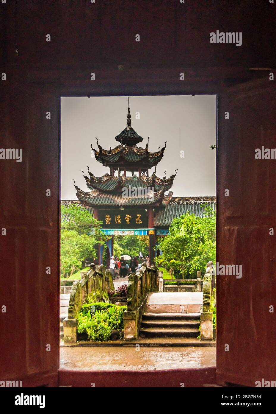 Fengdu, China - 8. Mai 2010: Geisterstadt, historisches Heiligtum. Blick durch braune Tür zu grünen Garten mit chinesischen architektonischen 3-Ebenen-Dach pag Stockfoto