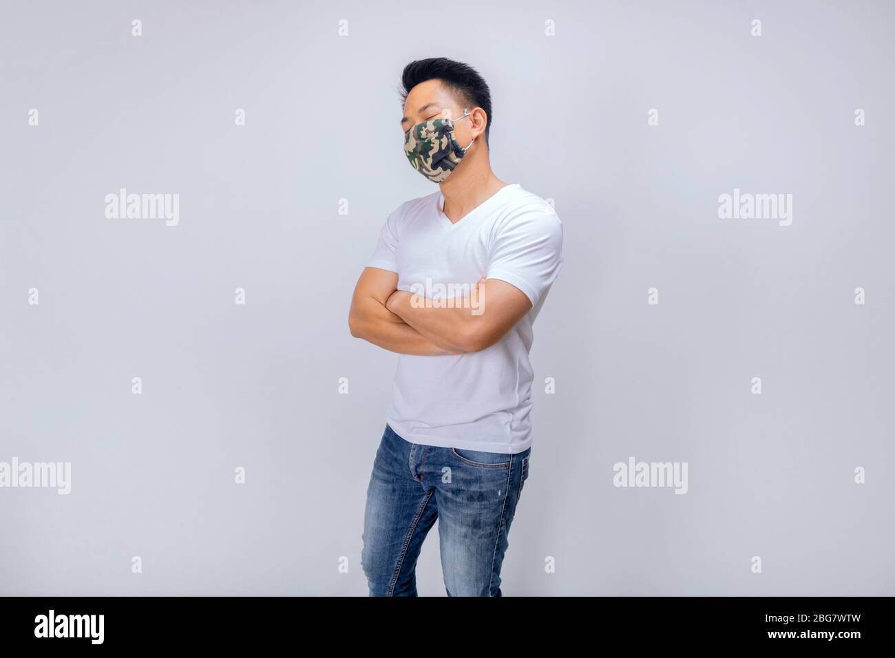 Ein asiatischer Kurzhaarmann in weißer Casual und Jeans posiert vor der Kamera in der Studiobeleuchtung, während er Tarnmaske trägt Stockfoto