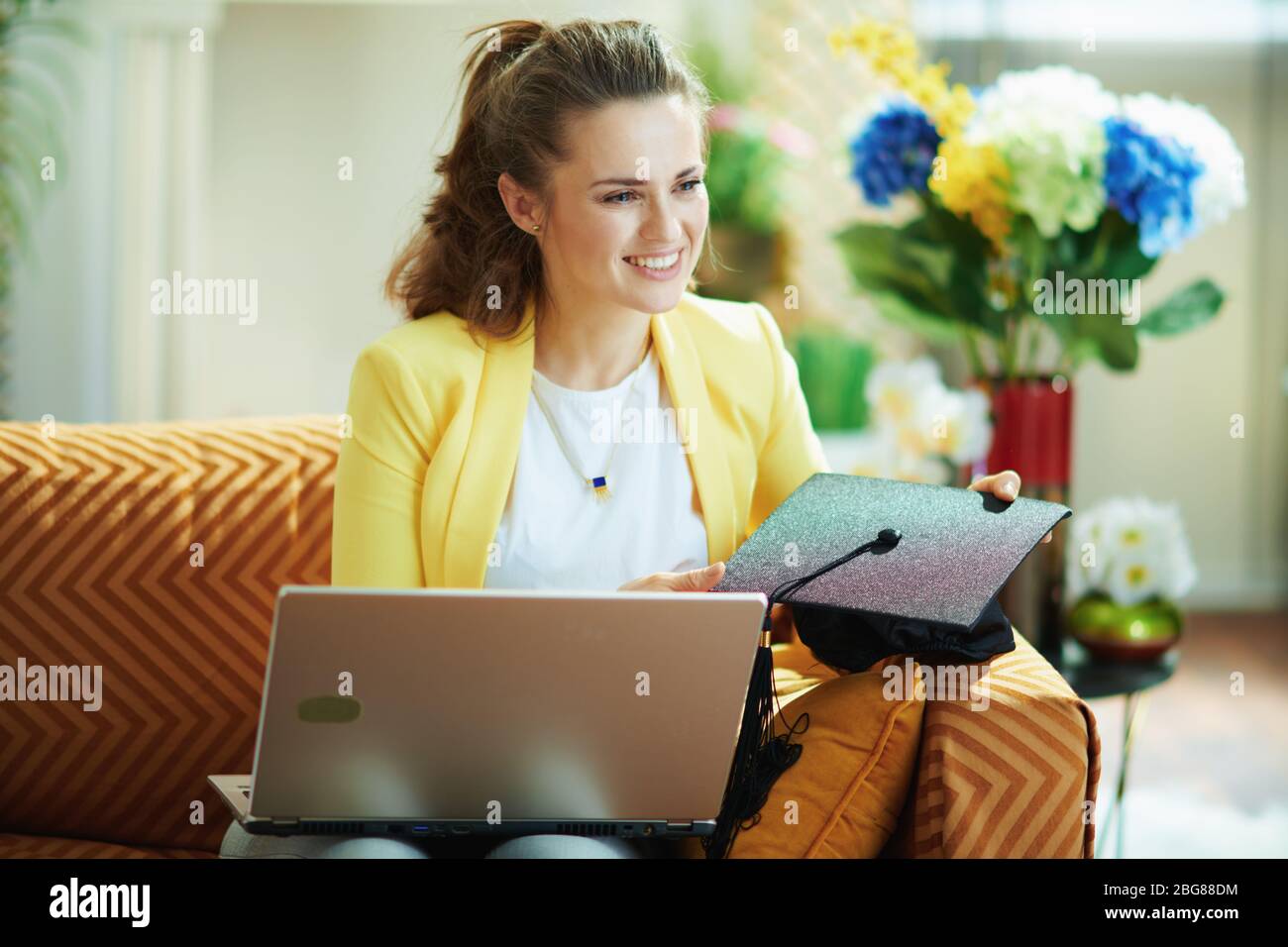 Nachdenkliche moderne Studentin in Jeans und gelber Jacke mit Abschlusskappe und Laptop im modernen Zuhause an sonnigen Tagen. Stockfoto