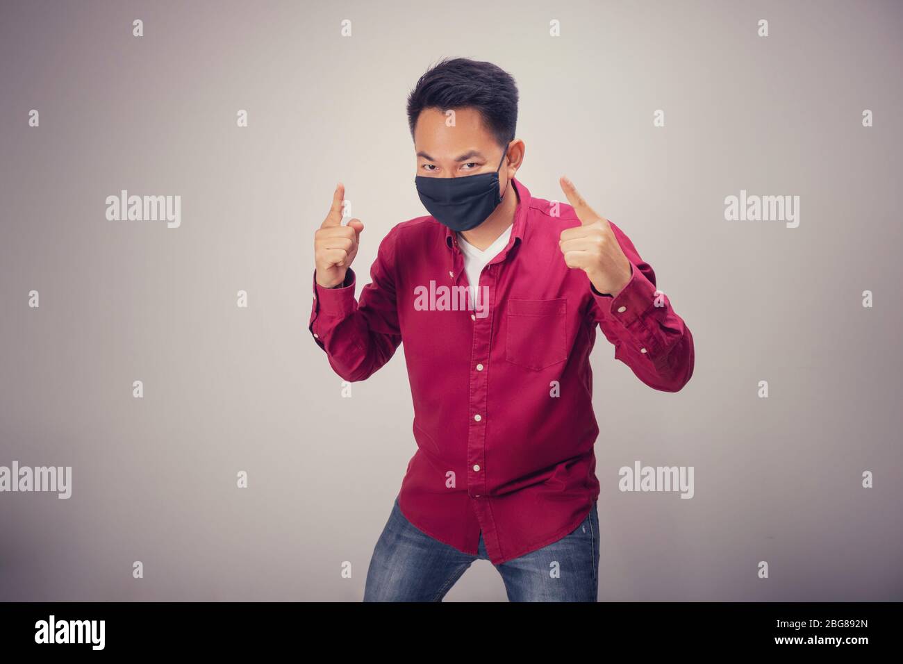 Ein asiatischer Kurzhaarmann in rotem Hemd zeigt in der Studiobeleuchtung mit schwarzer Maske den Finger Nummer eins vor der Kamera Stockfoto