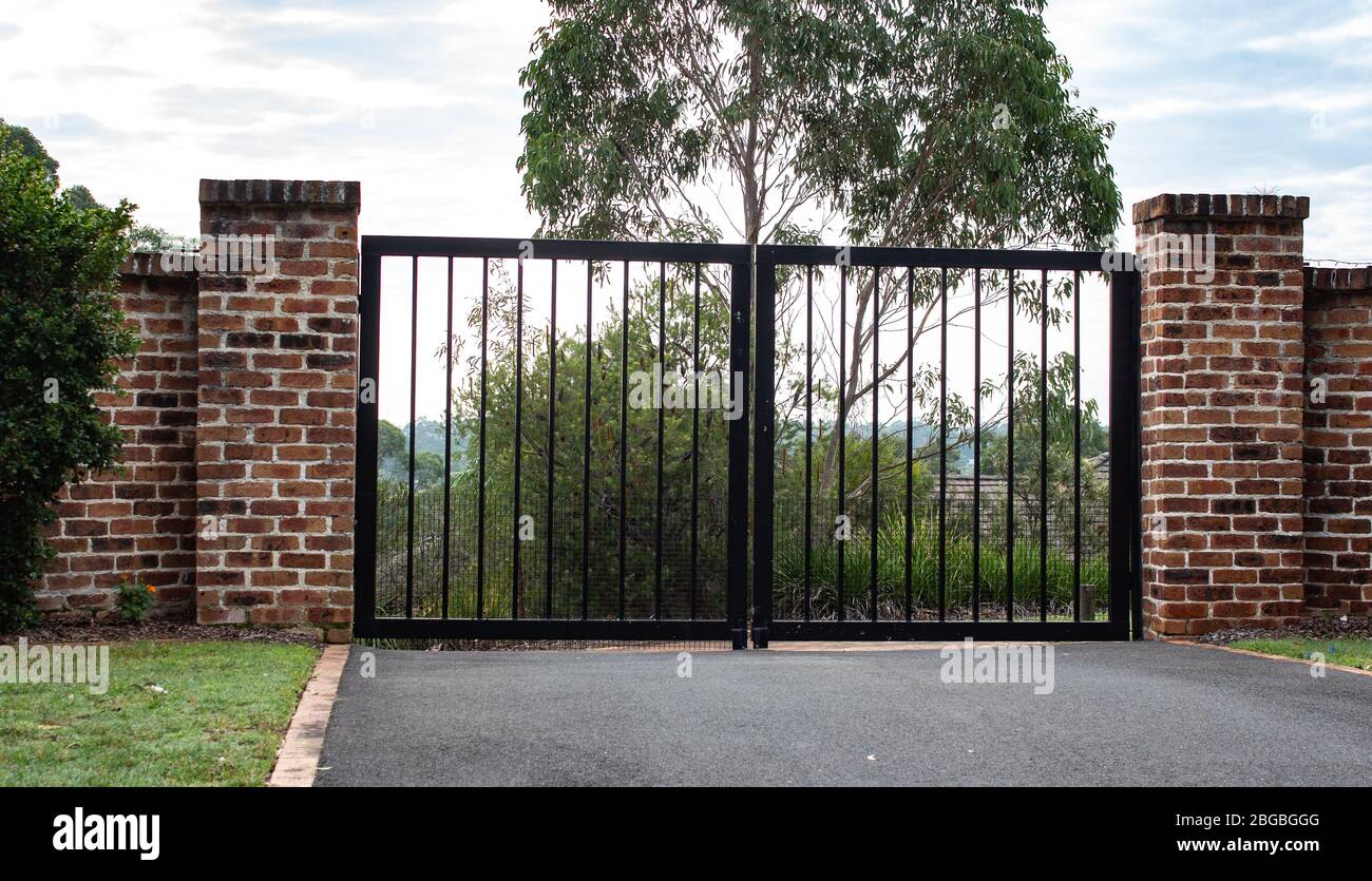 Einfahrtstore aus schmiedeeisernem schwarzen Metall, in Ziegelzaun, Lichter, grünem Gras, Gartenbäumen Stockfoto