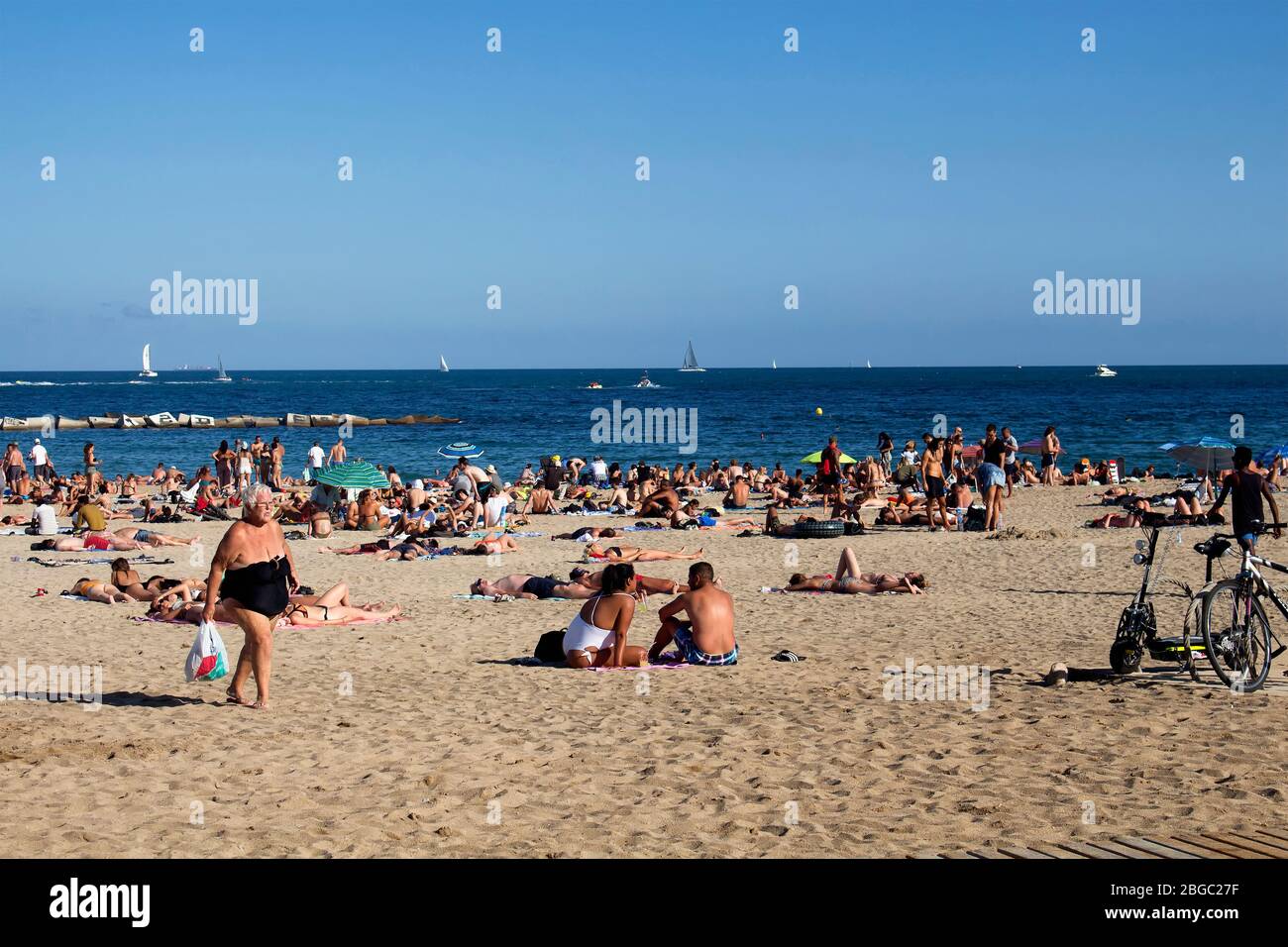 Blick auf viele Menschen, die sich auf Sand legen, sich sonnen und alte Frau am berühmten Strand 'La Barceloneta' in Barcelona spazieren gehen. Es ist ein sonniger Sumpfbecken Stockfoto