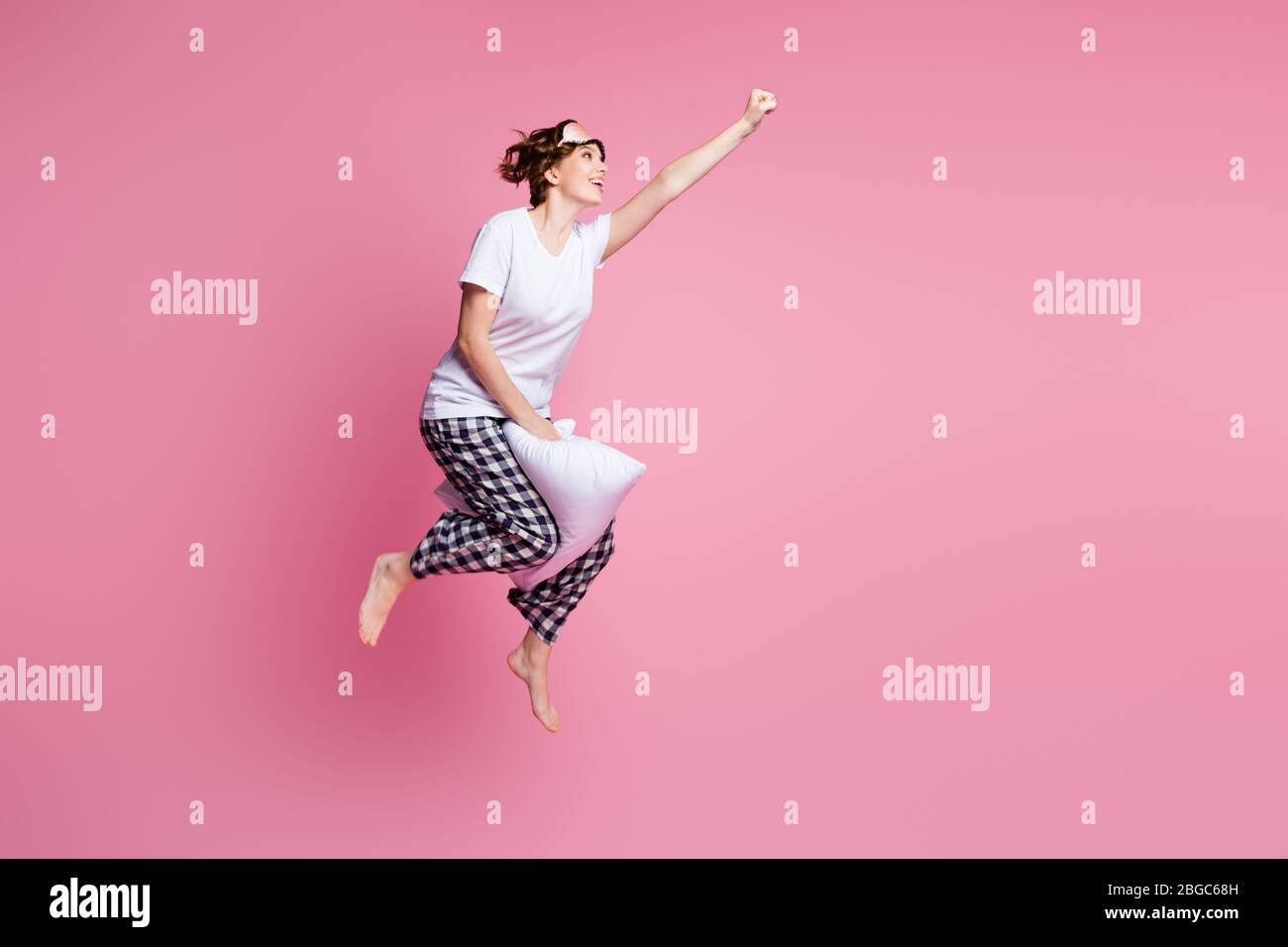 In voller Länge Foto von lustigen Dame springen hoch weichen großen Kissen zwischen den Beinen voran heben Faust tragen Maske weißen T-Shirt karierten Pyjama Hose barfuß Stockfoto