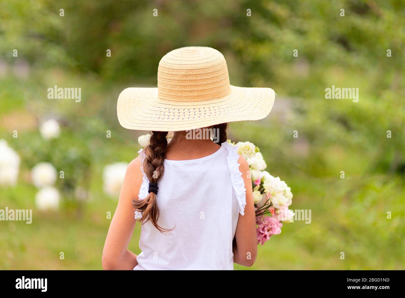 Ein Mädchen mit Zöpfen hält in ihren Händen einen Strauß weißer und rosa Pfingstrosen. Ein Kind in einem breitrandigen Strohhut an einem sonnigen Sommertag. Grüne Rückseite Stockfoto