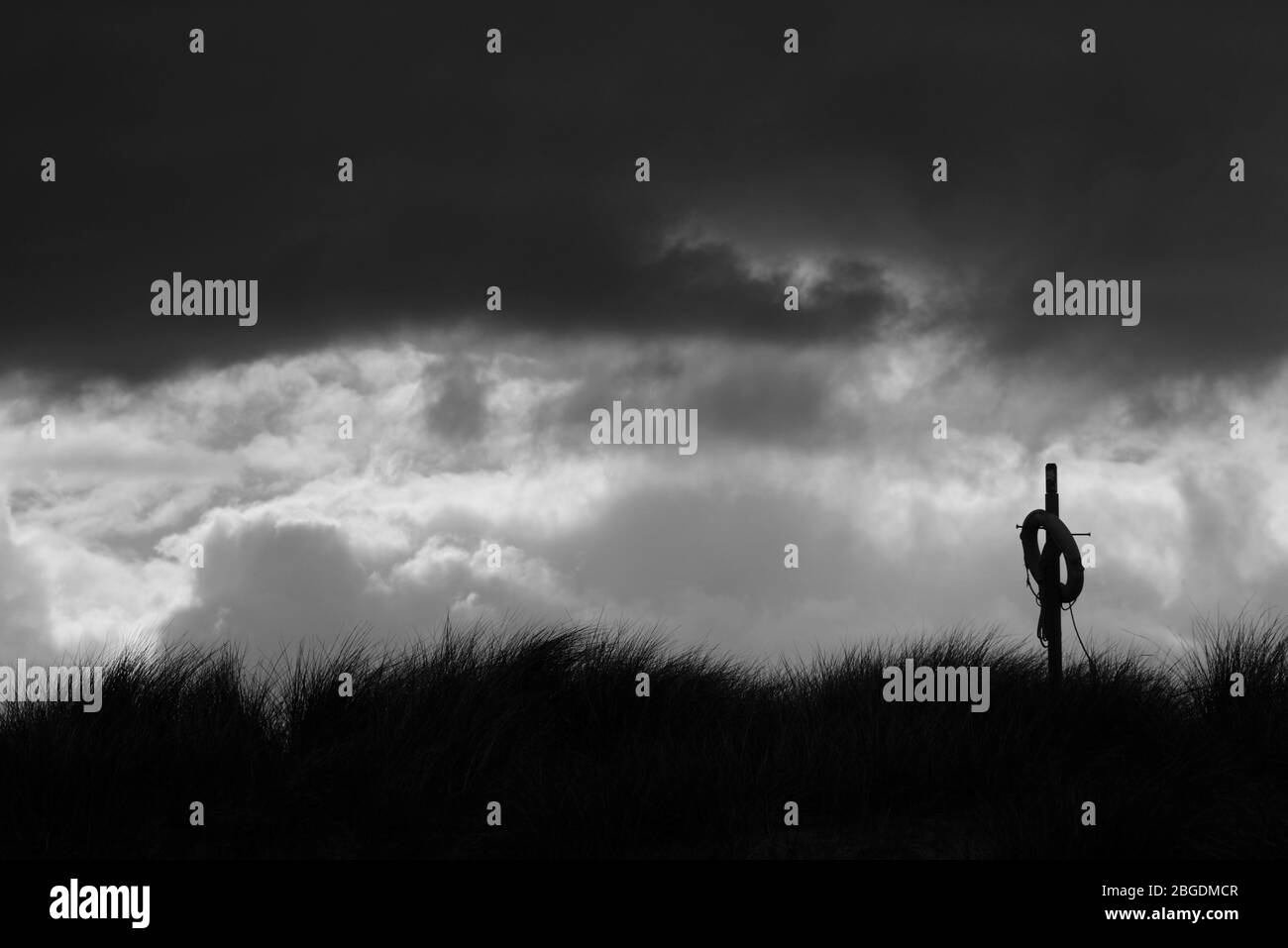Monochromes Bild der lebensrettenden Boje auf leicht zugänglichen Pfosten auf der Küstensanddüne in Silhouette und mit stürmischem Himmel dahinter Stockfoto