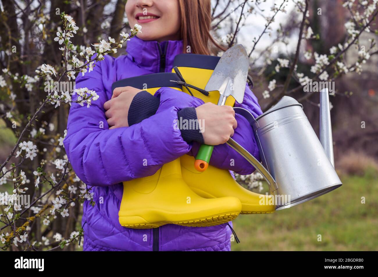 Ein Mädchen in einer violetten Jacke hält leuchtend gelbe Gummistiefel,  eine Gießkanne, einen Spaten in ihren Händen. Die Blütezeit der Bäume in  den Gärten und die Arbeitszeit auf persönlichen Parzellen. Frühling  Stockfotografie -