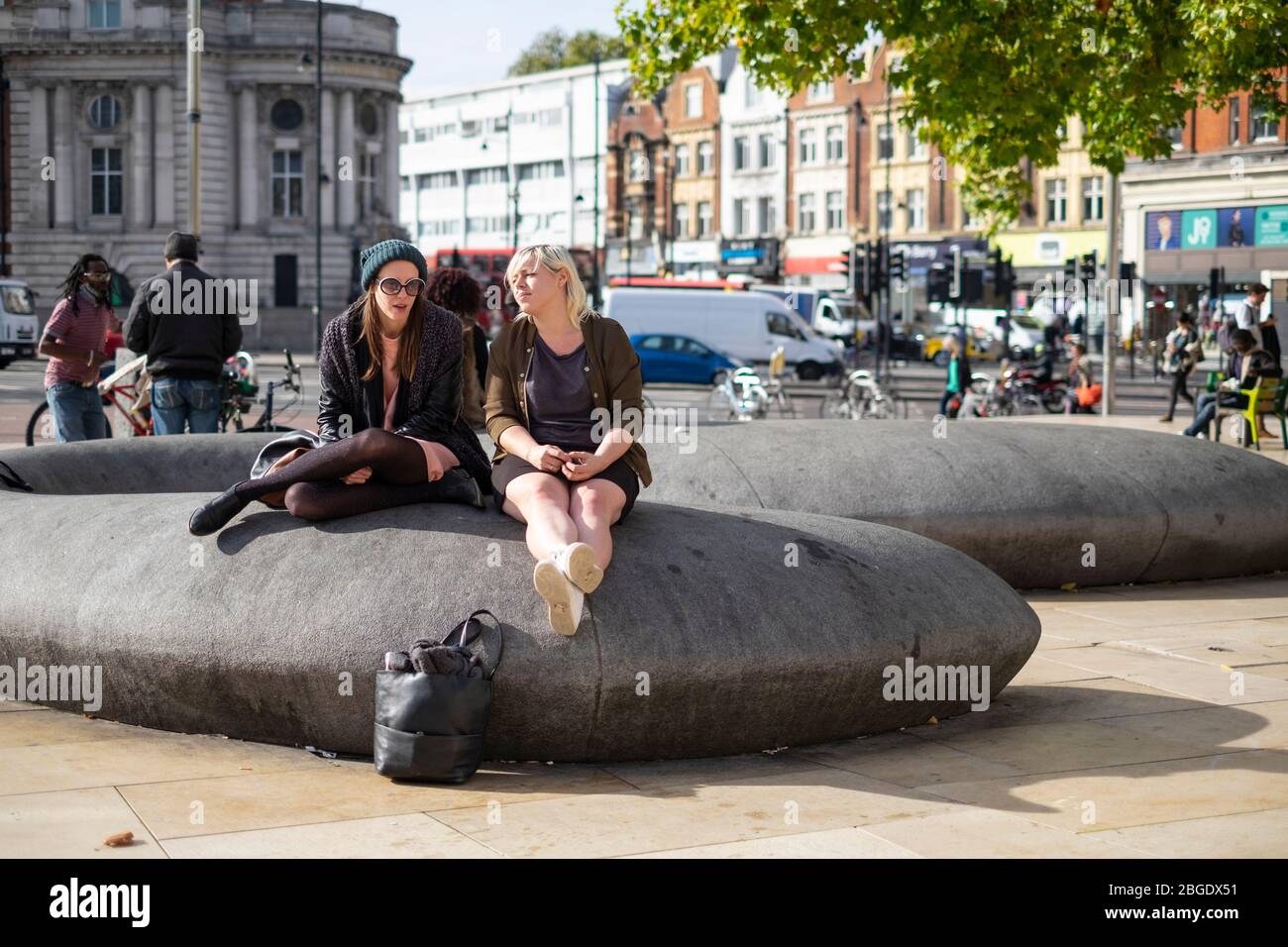 Zwei Mädchen sitzen auf einer Skulptur auf dem Windrush Square in der Sonne, Brixton, London Stockfoto