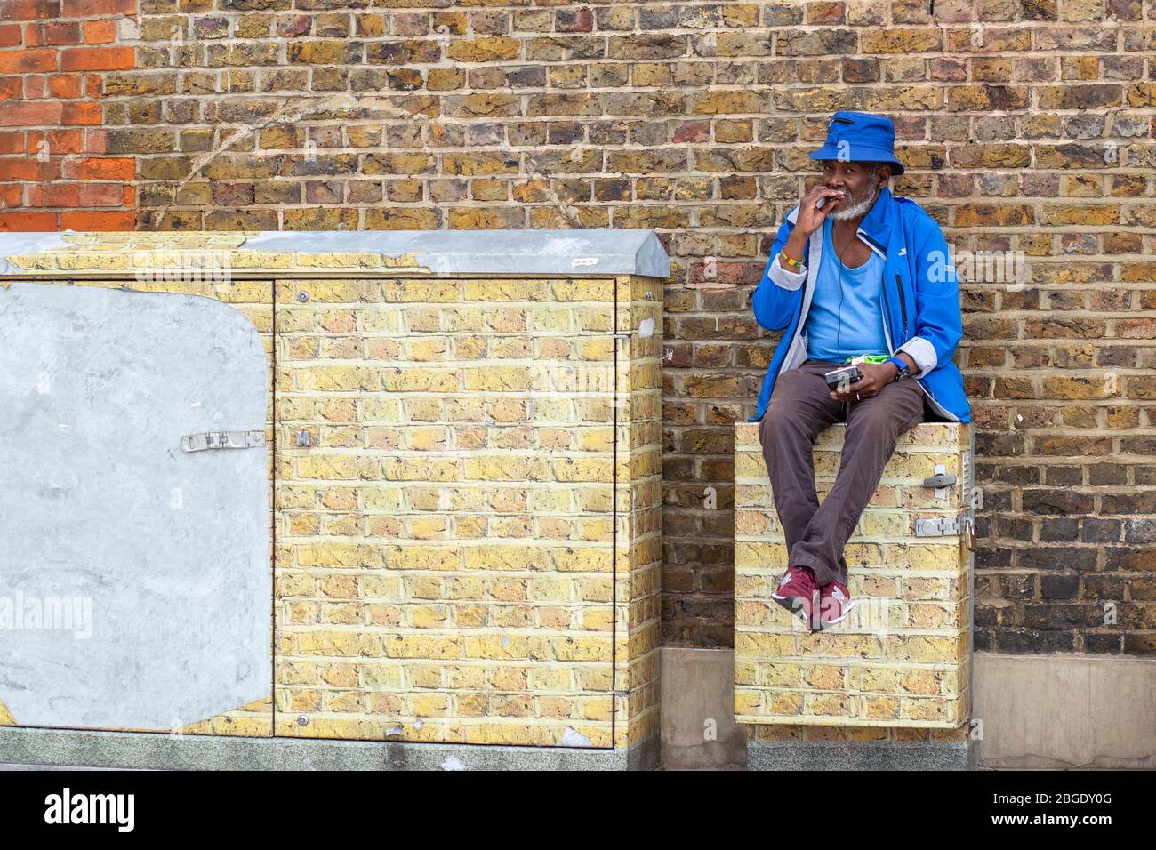 Ein Mann in einem blauen Mantel sitzt an einer Ziegelwand im Windrush Square, Brixton, London Stockfoto