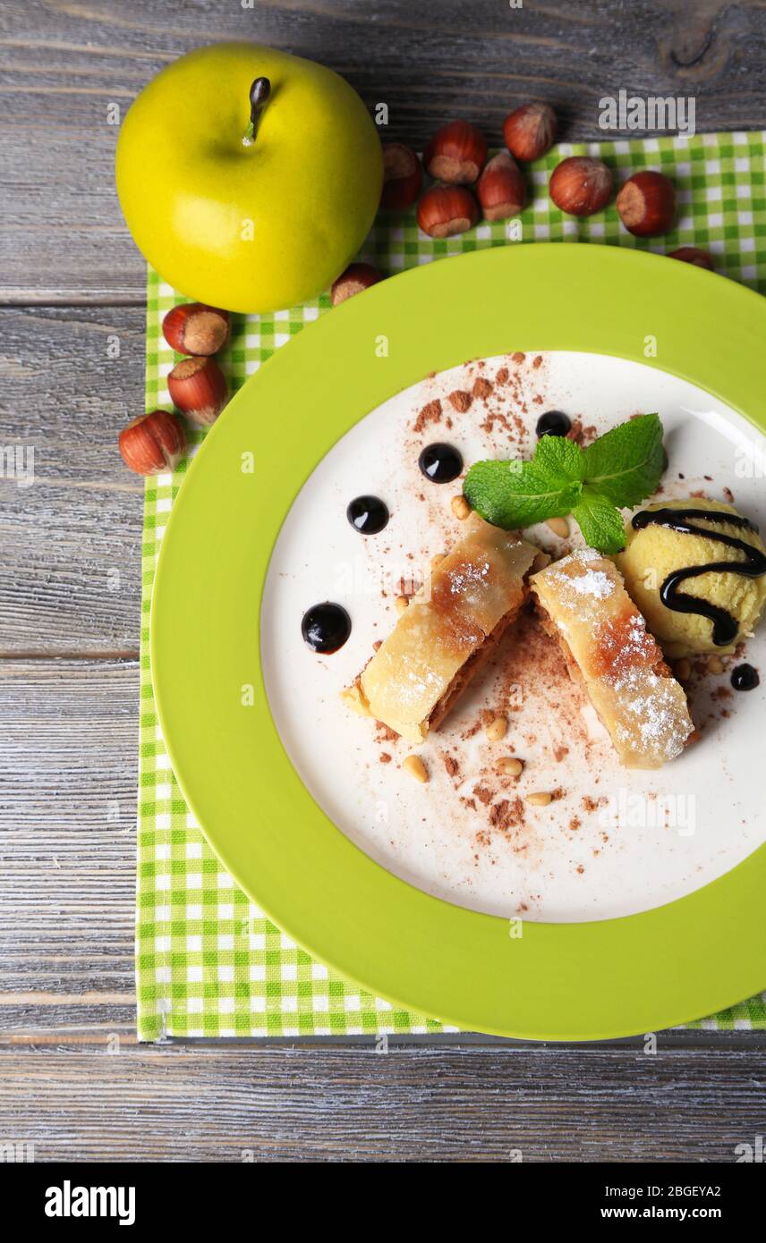 Leckerer selbstgemachter Apfelstrudel mit Nüssen, Minzblättern und Eis auf Teller, auf Holzhintergrund Stockfoto