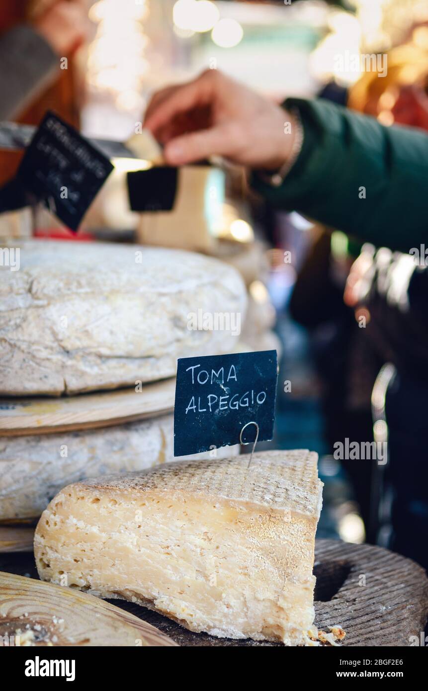 Marktstand mit italienischem Hartkäse in Scheiben geschnitten und offen, Alp-Käse-Etikett (Toma alpeggio) im Vordergrund und Menschen, die den Käse im Hintergrund kaufen Stockfoto