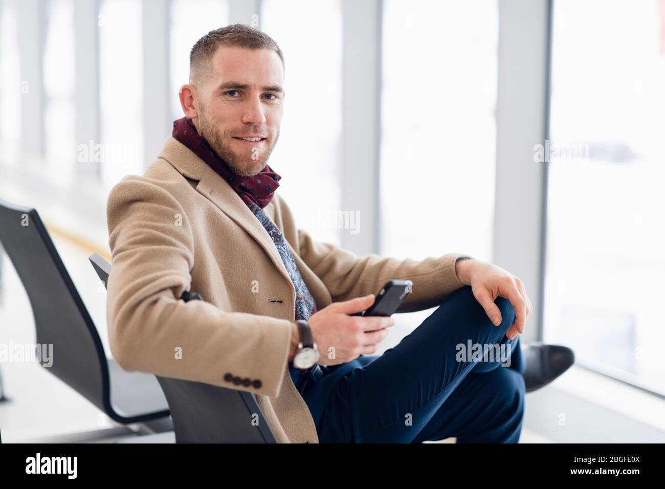 Attraktive junge Erwachsenen Mann in eleganter Schal und Sitzen im Flughafen Terminal Wartebereich Mantel. Er schaut auf zellulärer Mobiltelefon und Stockfoto