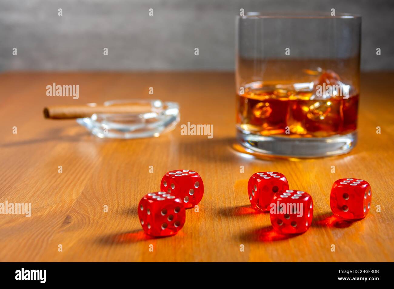 Rote Würfel auf einem Holztisch. Glas Whiskey mit Eiswürfeln und Aschenbecher mit Zigarre im Hintergrund Stockfoto
