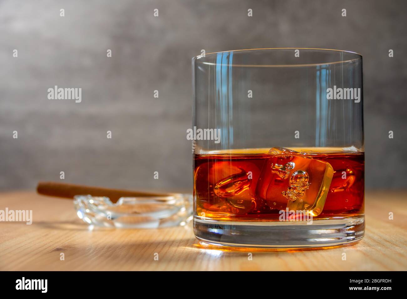 Glas Whiskey mit Eiswürfeln auf dem Holztisch. Ein Aschenbecher mit einer Zigarre im Hintergrund in Unschärfe-Effekte Stockfoto