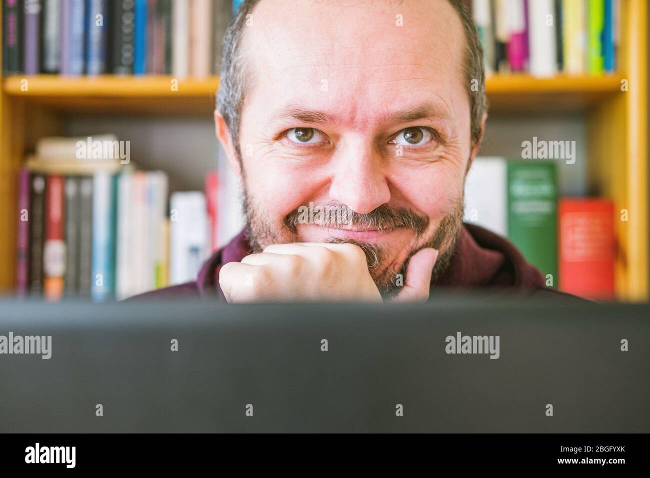 Erwachsener Mann arbeitet von zu Hause aus. Bärtiger Mann, der online von zu Hause aus auf einem Computer-Laptop arbeitet, Bücherregale hinter ihm. Gesichtsausdruck, Lächeln. Nahaufnahme Stockfoto