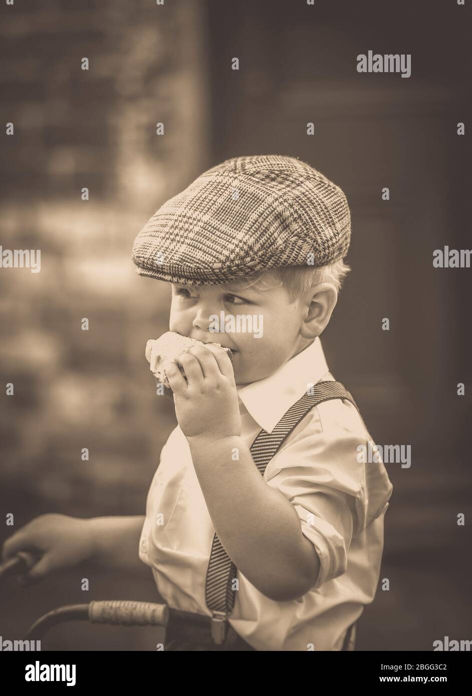 Vintage Sepia Nahaufnahme des süßen kleinen 1940er-Jungen mit flacher Mütze und Hosenhalterung mit dreirädrigem, isoliertem Sandwich im Freien, 1940er WW2. Event, UK. Stockfoto