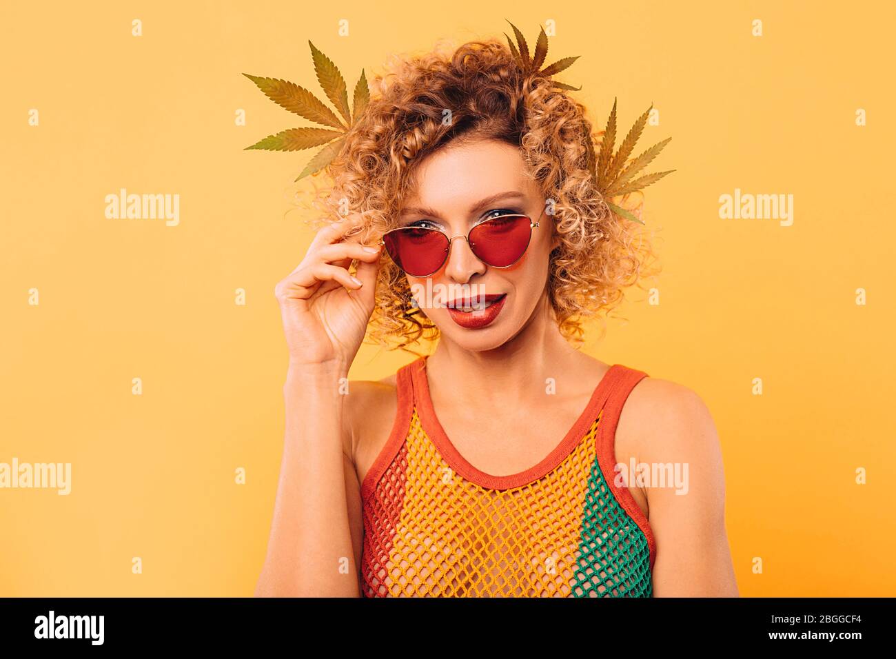 Lockige Frau in rosa Brille und einem Rastakleid mit Marihuanablättern in den Haaren auf gelbem Hintergrund Stockfoto