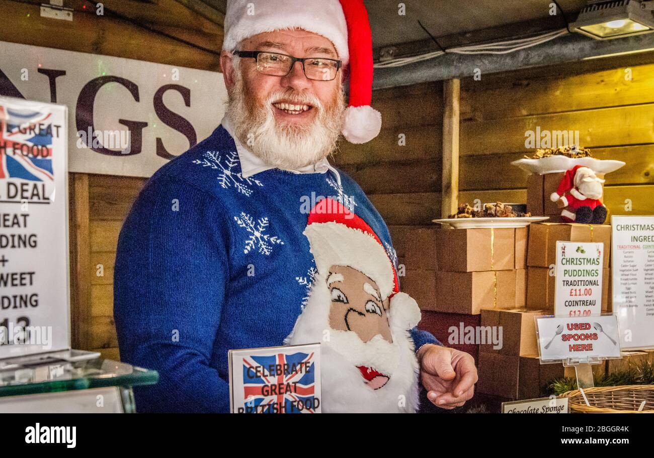Bärtiger, lächelnder Stallbesitzer, der als Weihnachtsmann gekleidet ist und auf der Waddesdon Manor Food Fair - 2016 Weihnachtspudding verkauft Stockfoto