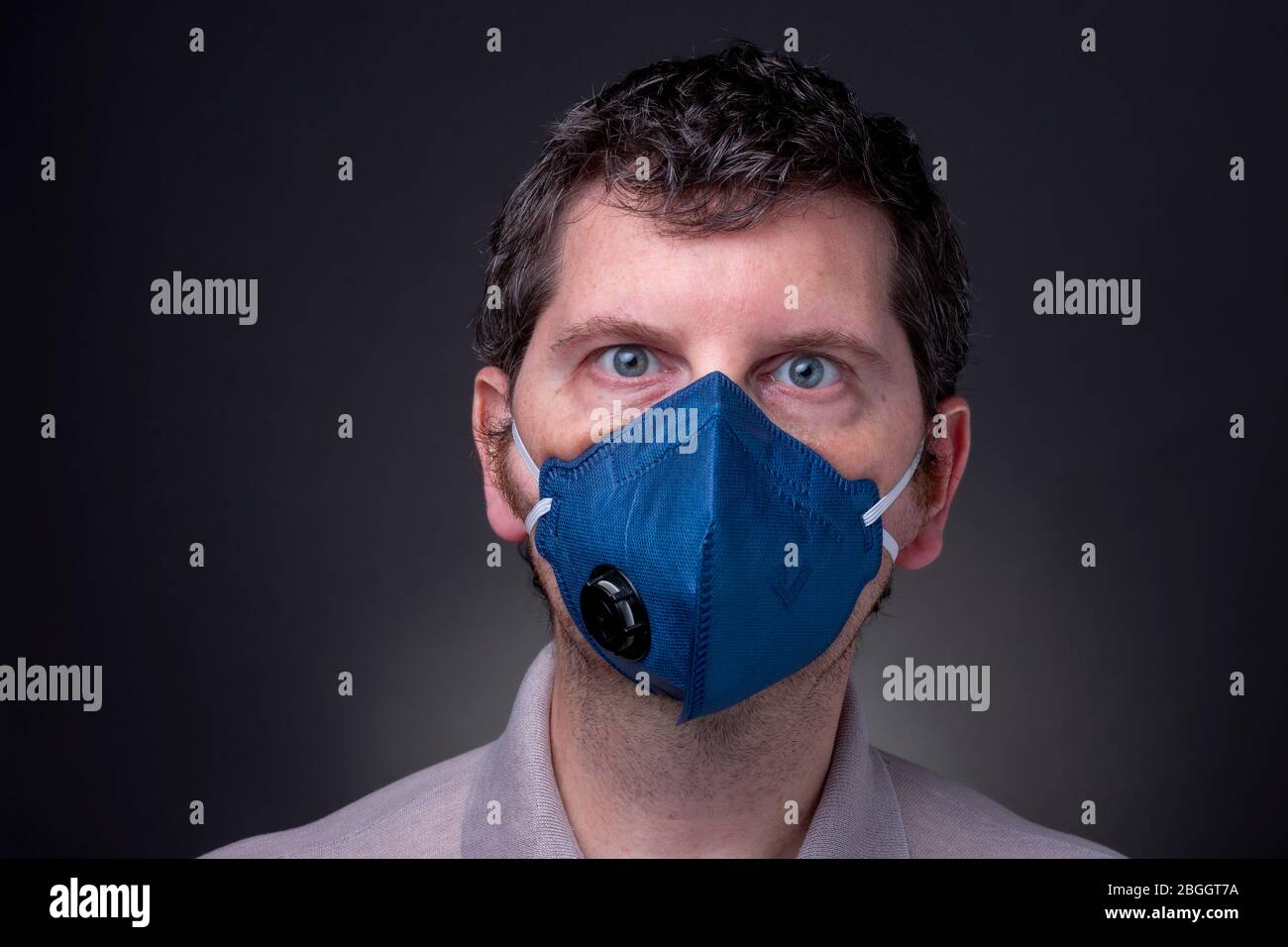 Kaukasischer Rüde mit dunkelblauer N95 Atemschutzmaske. Studio Low-Key-Profil des Mannes mit weit offenen stressigen blauen Augen und verwirrten Blick Stockfoto
