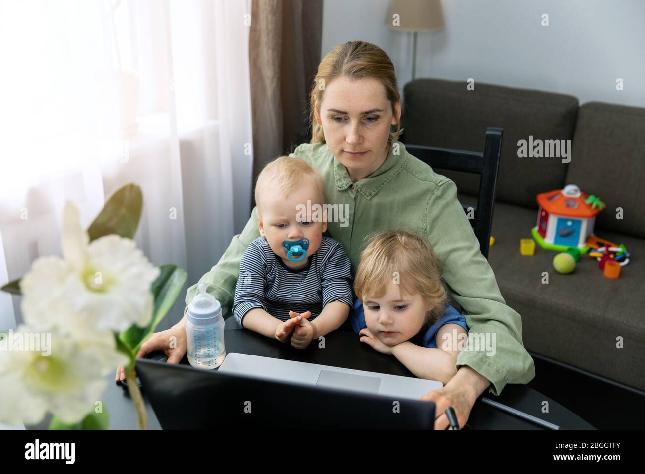 Junge Frau, die während der Quarantäne von zu Hause aus arbeitet. Laptop am Schreibtisch mit zwei Kindern auf dem Schoß Stockfoto