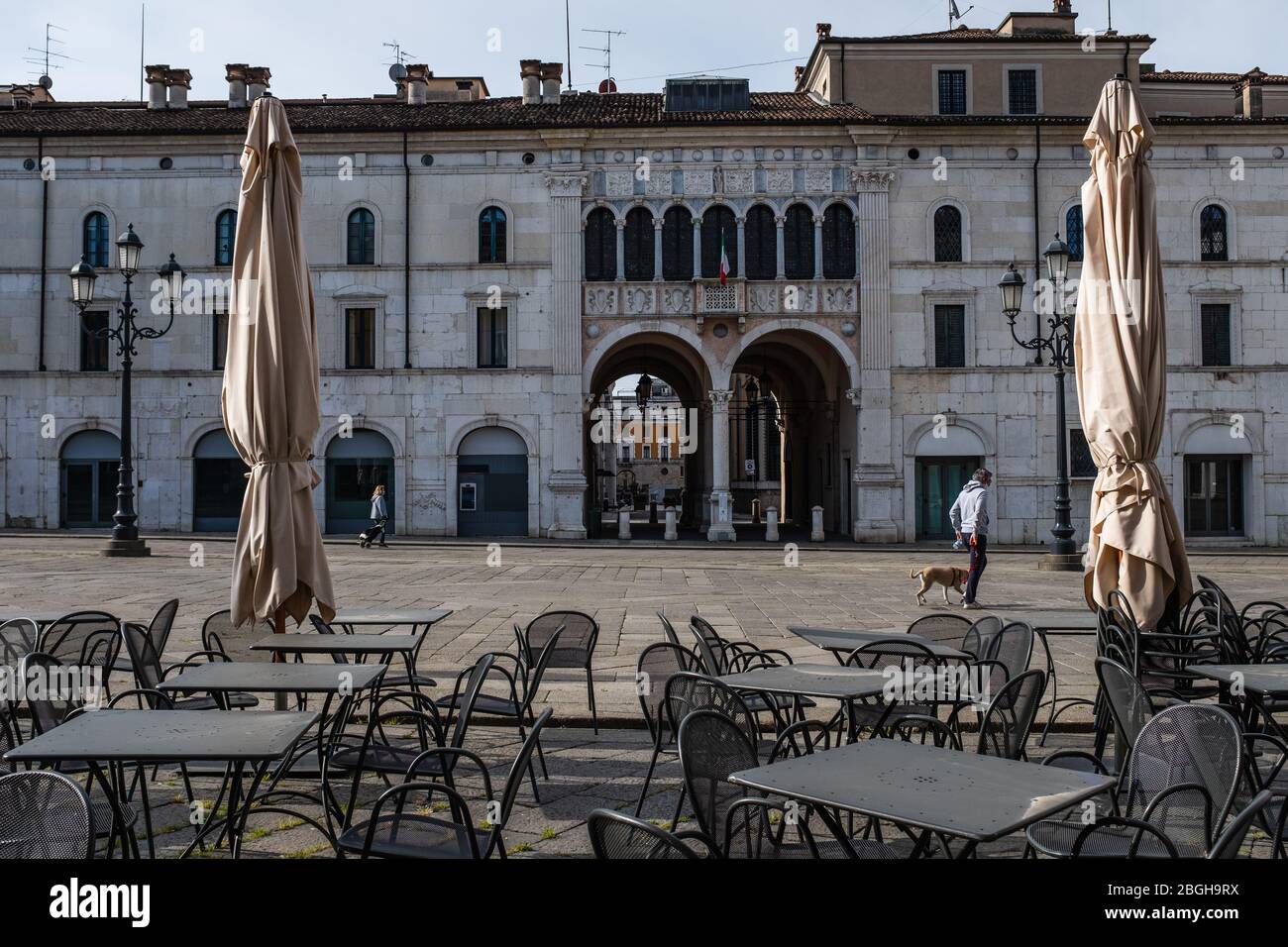 Zwei Leute gingen über den Hauptplatz in Brescia (Italien). Menschenleere Tische vor einem Restaurant während der Coronavirus-Notabsperrung. Stockfoto