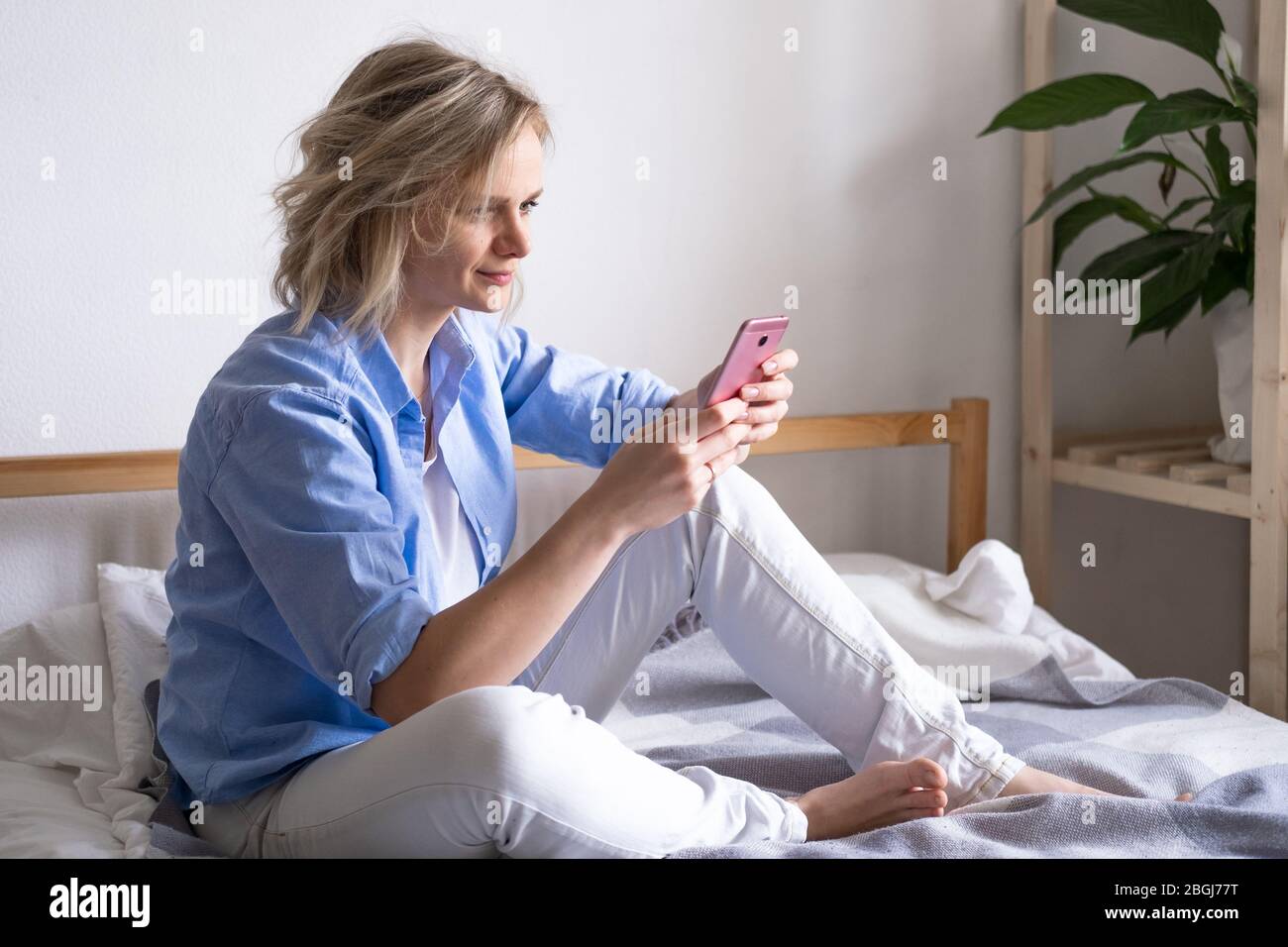 Blonde Frau mit Smartphone auf dem Bett sitzen. Lesen von Nachrichten und Online-Büchern. Soziale Medien, instagram, zu Hause bleiben und Quarantäne Stockfoto