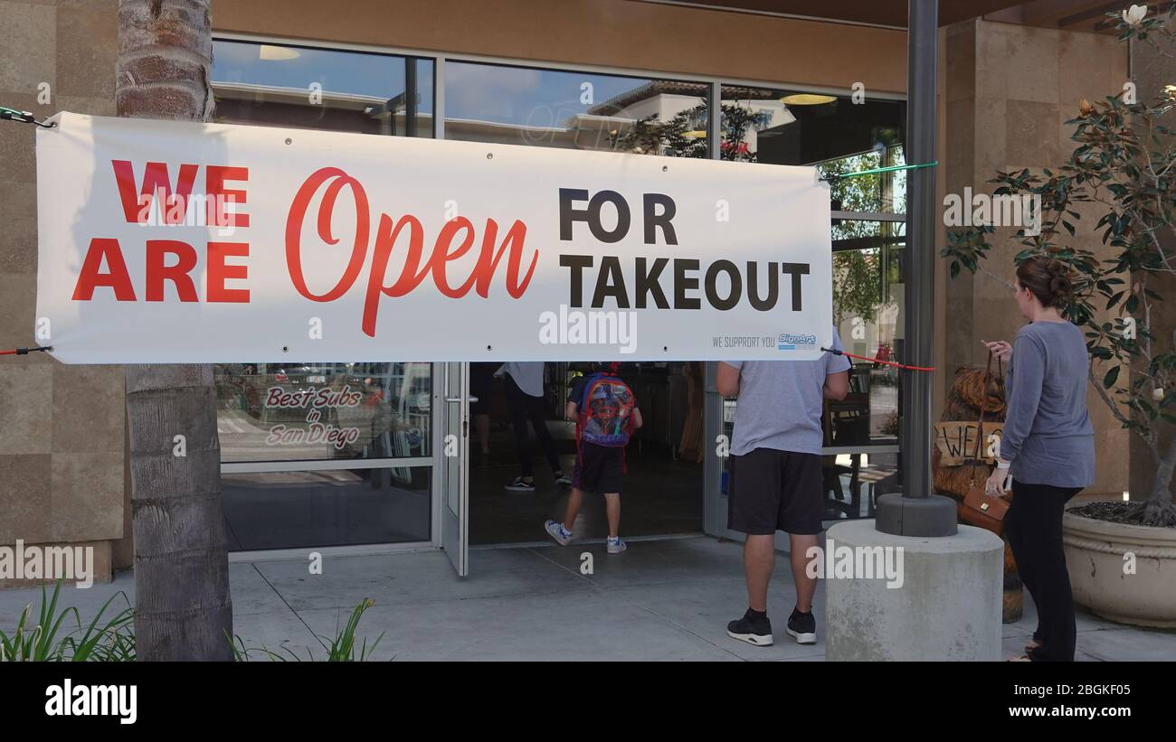 Ein großes Schild an einem Deli-Laden erinnert die Leute daran, dass sie für Essen zum Mitnehmen geöffnet sind. Menschen in der Linie üben soziale Distanz. Stockfoto