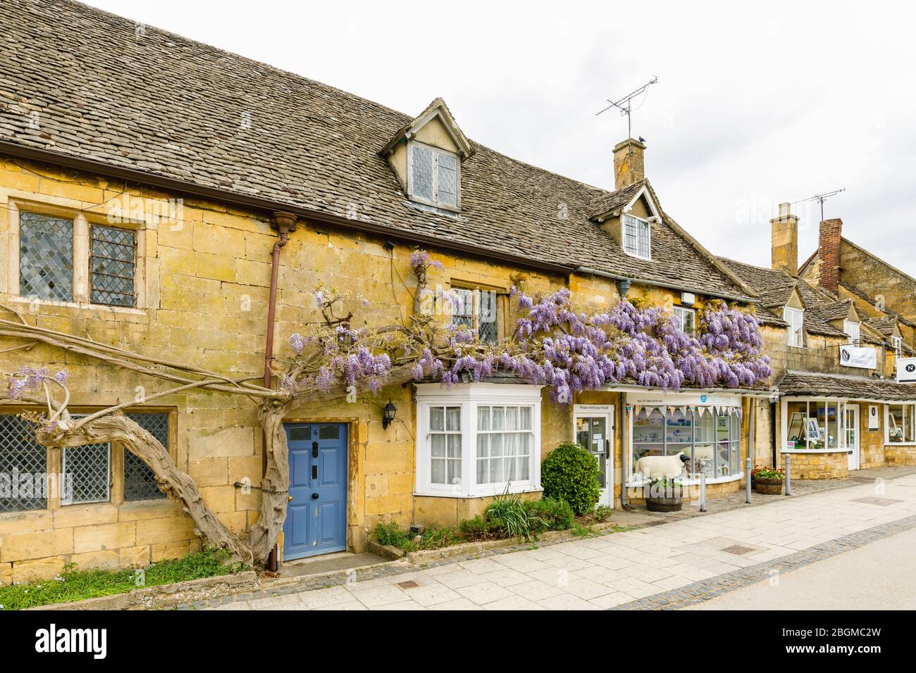 Geschäfte am Straßenrand, lokaler Stil und Glyzinien in Blumen in High Street, Broadway, Worcestershire, ein wunderschönes Dorf in den Cotswolds, Südwestengland Stockfoto
