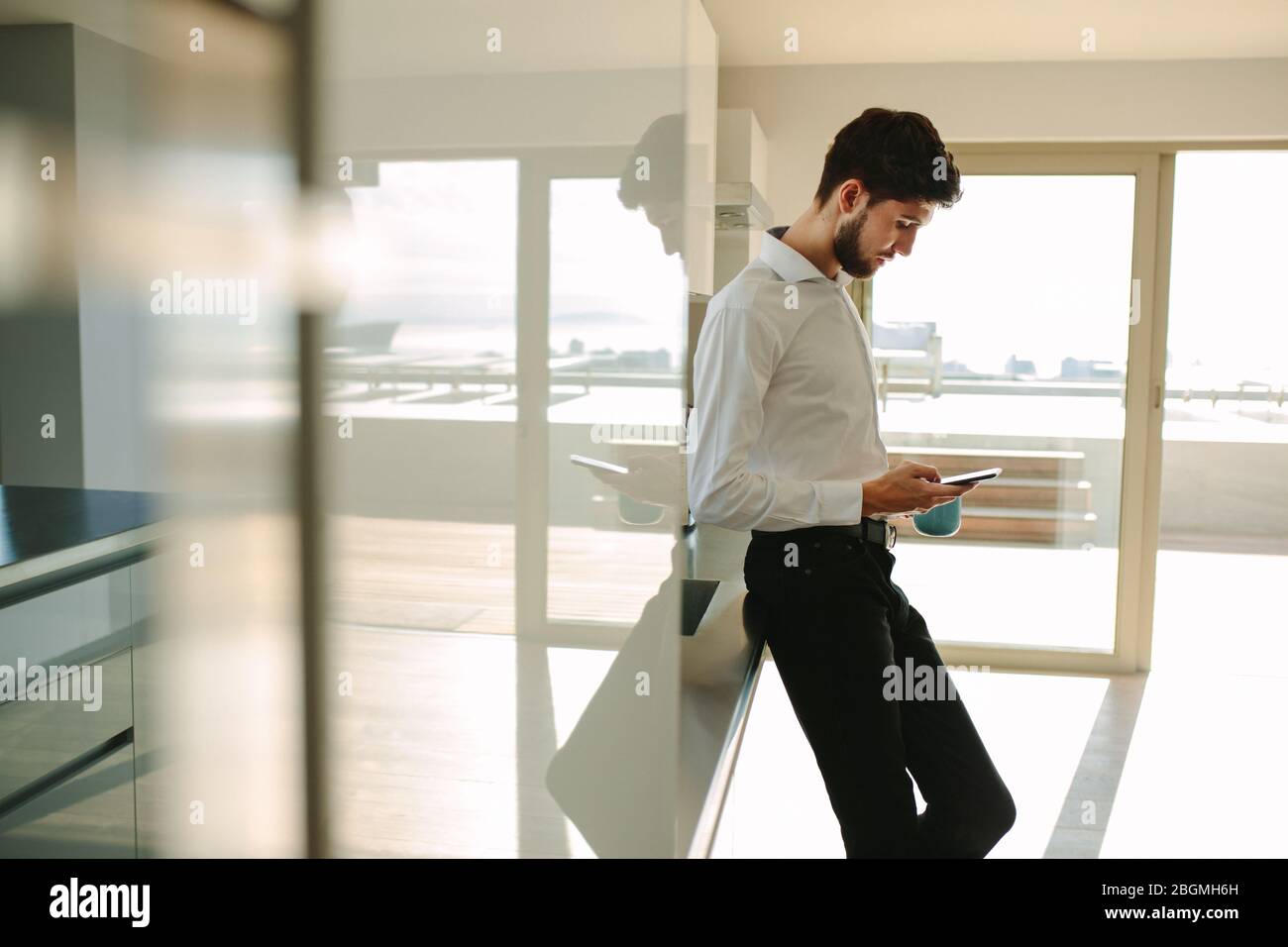 Mann, der in der Küche steht und eine Tasse Kaffee hält und das Handy benutzt. Geschäftsmann, der Smartphone sieht, während er eine Tasse Kaffee in der Küche hält Stockfoto