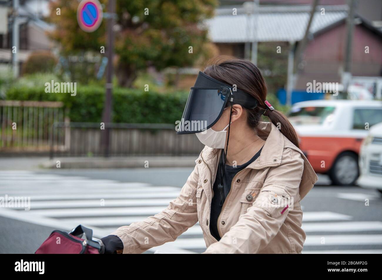 Kyoto, Japan - 12. April 2019: Junge Radfahrerin auf einer belebten Straße, die eine Anti-Umweltverschmutzung-Gesichtsmaske und ein dunkles Visier trägt, um sich vor der Sonne zu schützen. Stockfoto