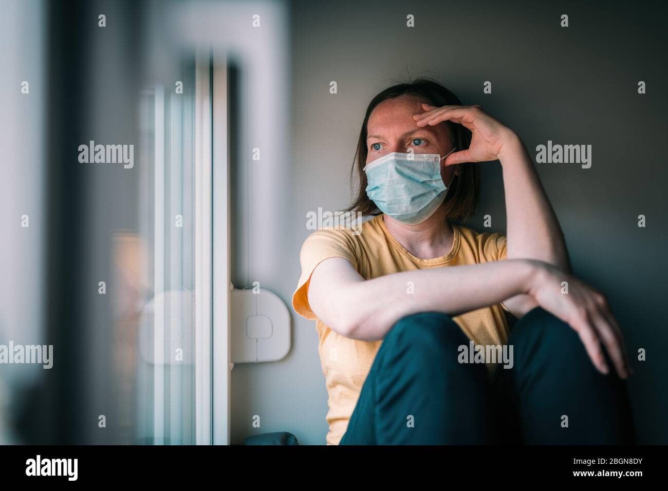 Frau in Selbstisolation während des Virenausbruchs, die durch das Fenster schaut. Besorgte weibliche Person mit schützenden chirurgischen Maske in zu Hause bleiben Konzept, sel Stockfoto