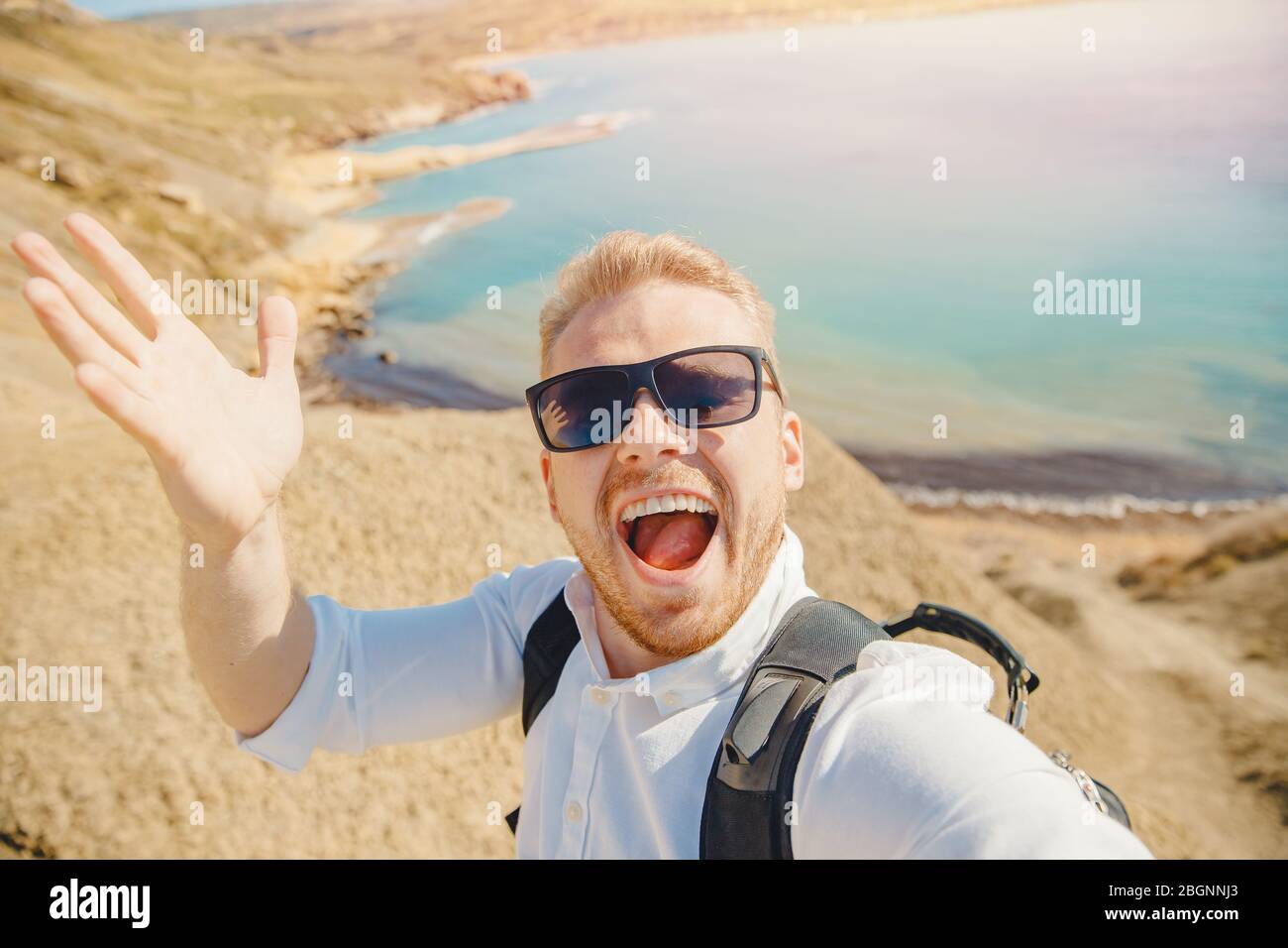 Männliche Reisende macht Selfie-Foto auf Hintergrund des blauen Meeres, Sandstrand in Sonnenbrille und mit Rucksack. Reisekonzept Stockfoto