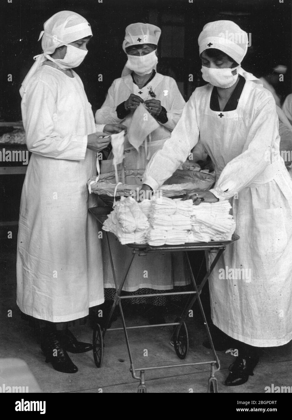 Zum Schutz vor der spanischen Grippe maskiert, entfernen die Arbeiter des Roten Kreuzes Bündel von Masken für amerikanische Soldaten vom Tisch, wo andere Frauen eifrig daran beteiligt sind, sie zu machen. New York, 1918. Stockfoto