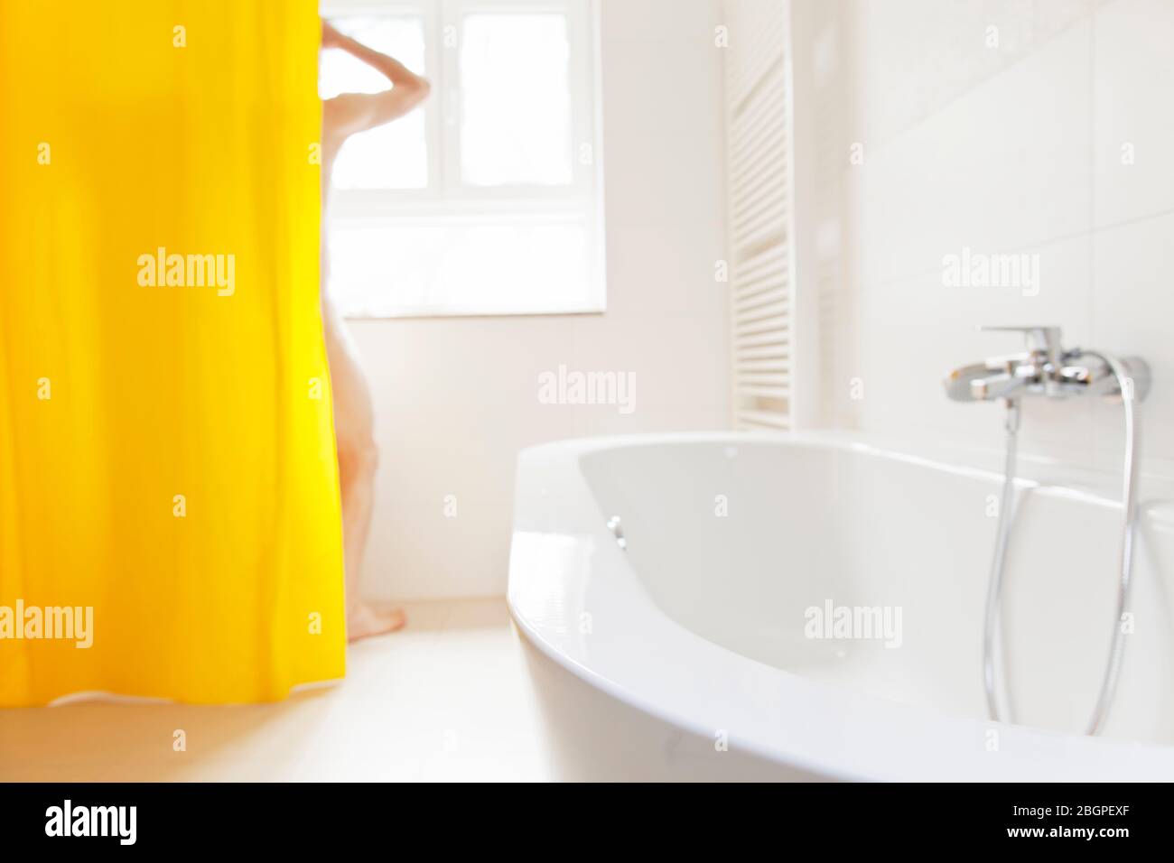 Mann, der in einem hellen Badezimmer mit gelbem Duschvorhang duscht - Fokus auf die Badewanne im Vordergrund Stockfoto
