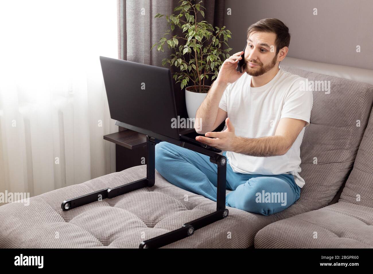 Telefongespräch. Stress kaukasischen düsteren Mann sprechen Telefon arbeiten aus dem Home-Office mit Laptop und Internet. Remote-Arbeit im Stress, Freiberufler Stockfoto