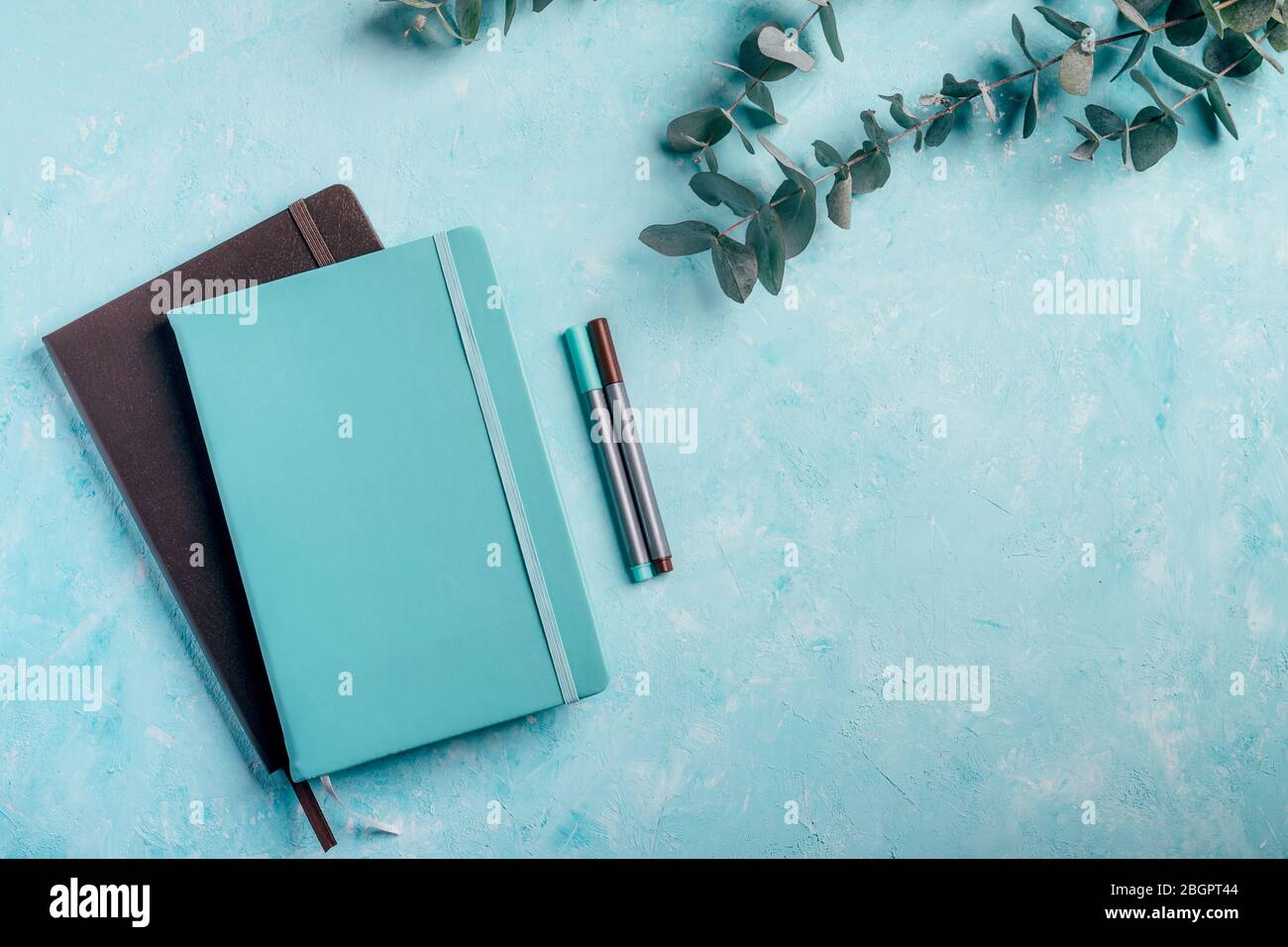 Draufsicht auf blaue und braune Notizbücher und Fineliner-Marker mit grünen Eukalyptuspflanzen auf türkisfarbenem, rustikalem Schreibtischhintergrund. Stockfoto