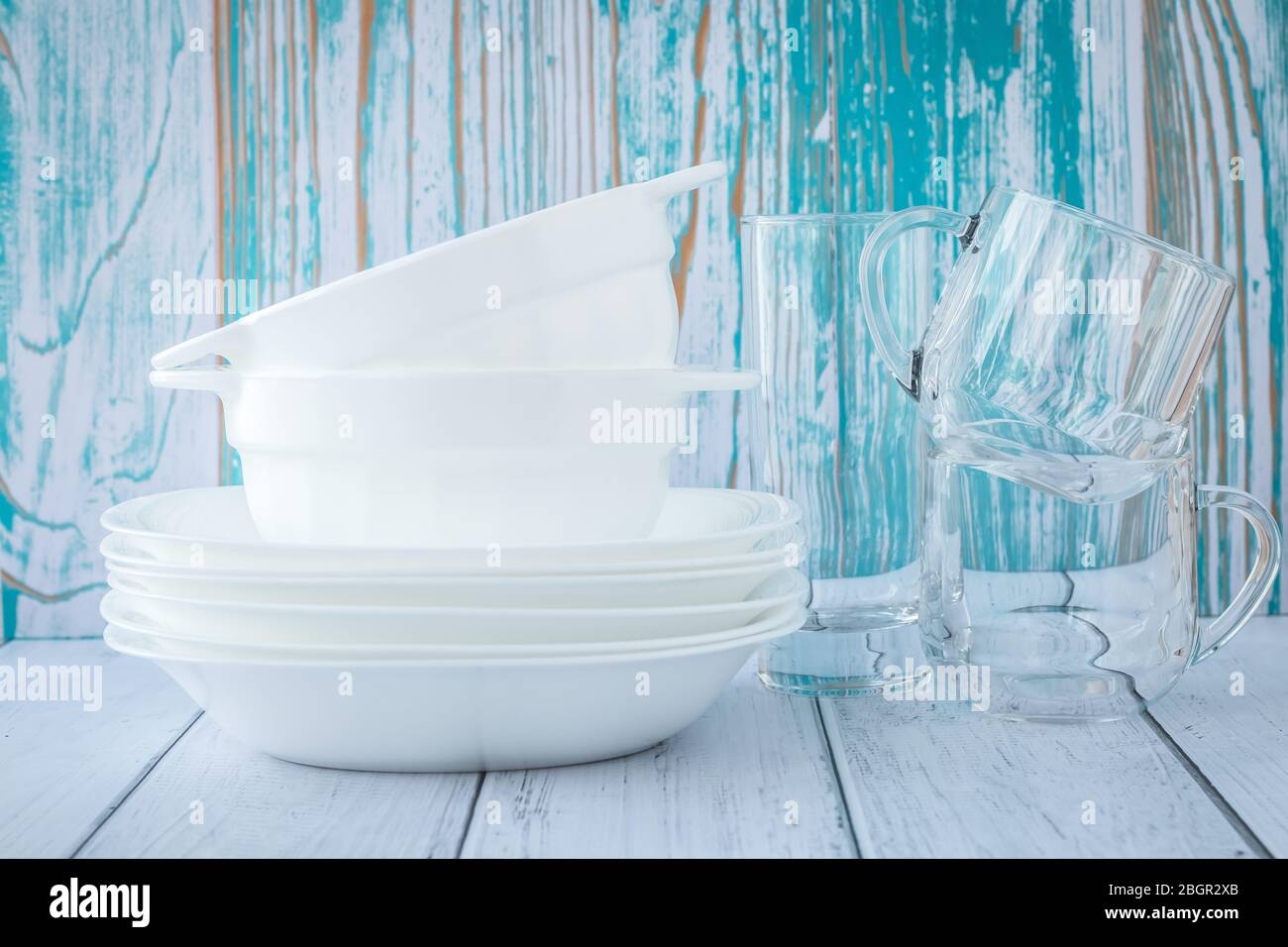 Ein Stapel von sauberen Geschirr auf einem türkisfarbenen Holzhintergrund. Weiße Keramikplatten und transparente Gläser Stockfoto