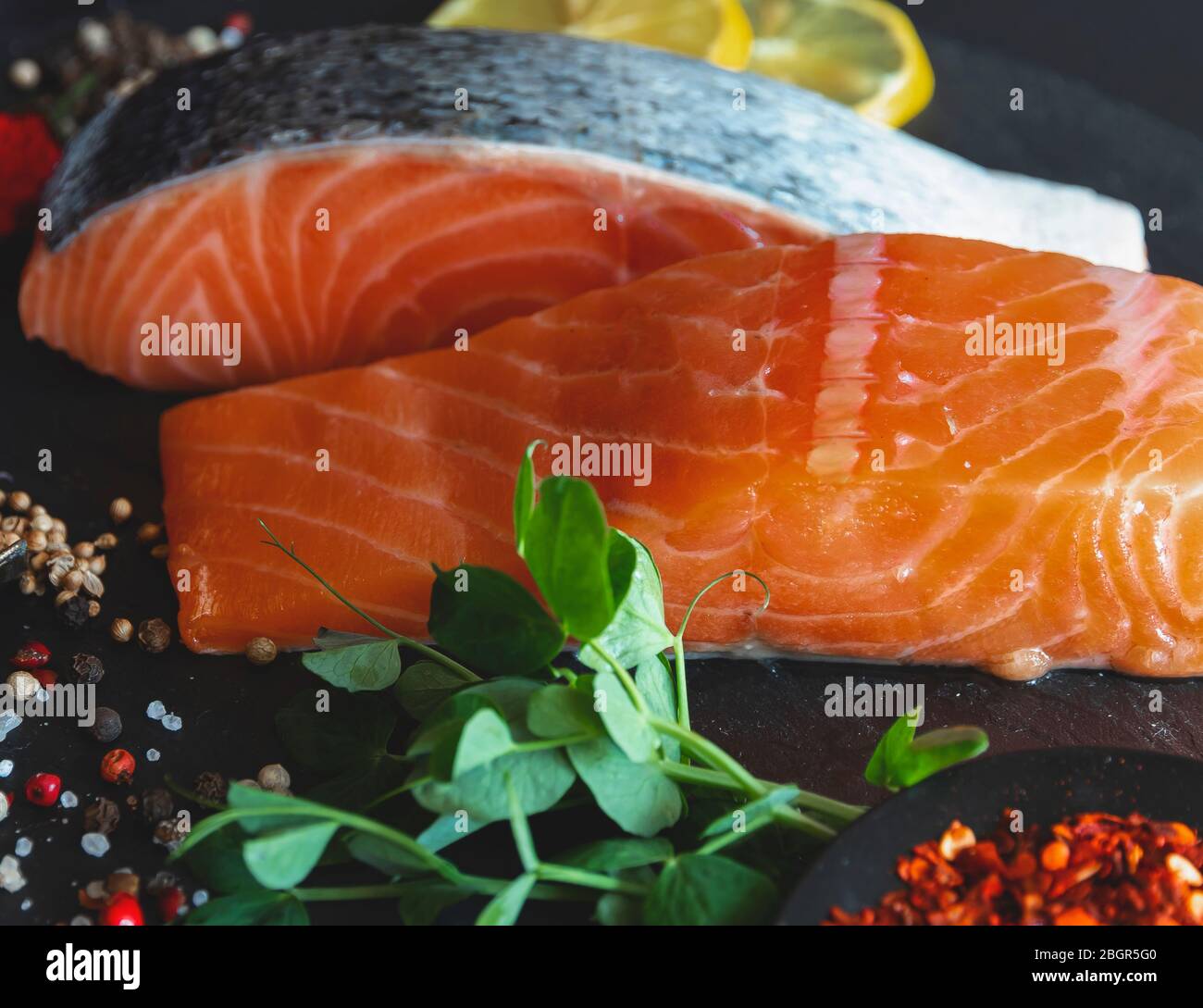 Stücke von rohem Lachsfischfilet, Gewürzen und Erbsensprossen liegen auf einer Steinoberfläche Stockfoto