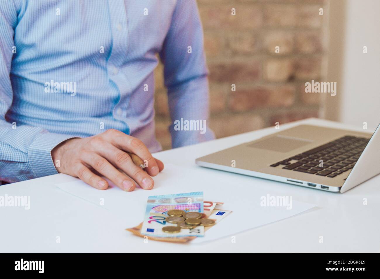 Mann, der an einem Schreibtisch mit Laptop sitzt und Zeitungen in hellblauem Hemd liest. Leichter Arbeitsplatz, Geschäftsraum. Arbeitsumgebung. Backsteinmauer Hintergrund. Stockfoto