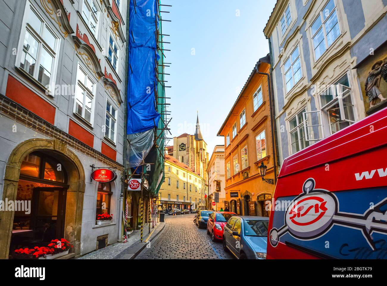Eine enge Straße mit Geschäften, gesäumt von Autos und Lastwagen in Prag, Tschechische Republik, während Sonnenlicht einen Kirchturm im Stadtteil Male Strana hervorhebt Stockfoto