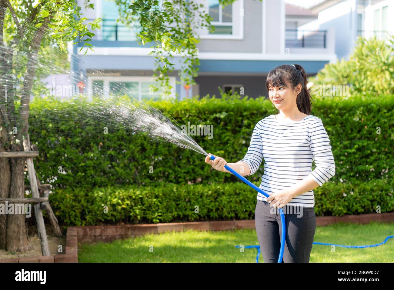 Asiatische Frau glücklich in Haus Hinterhof Sommergarten, mit Gartenschlauch Spritzwasser auf dem Rasen und Baum Blätter während des Aufenthalts zu Hause mit kostenlos Stockfoto