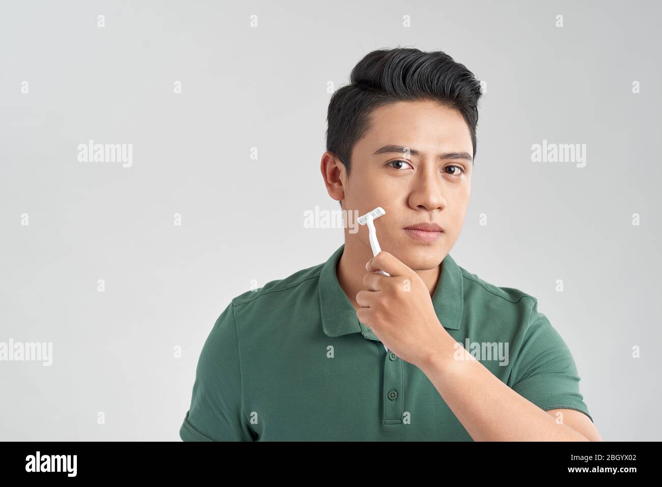 Schöner junger Mann, der sich sein Gesicht rasieren und die Kamera betrachten kann, während er isoliert auf grauem Hintergrund steht Stockfoto