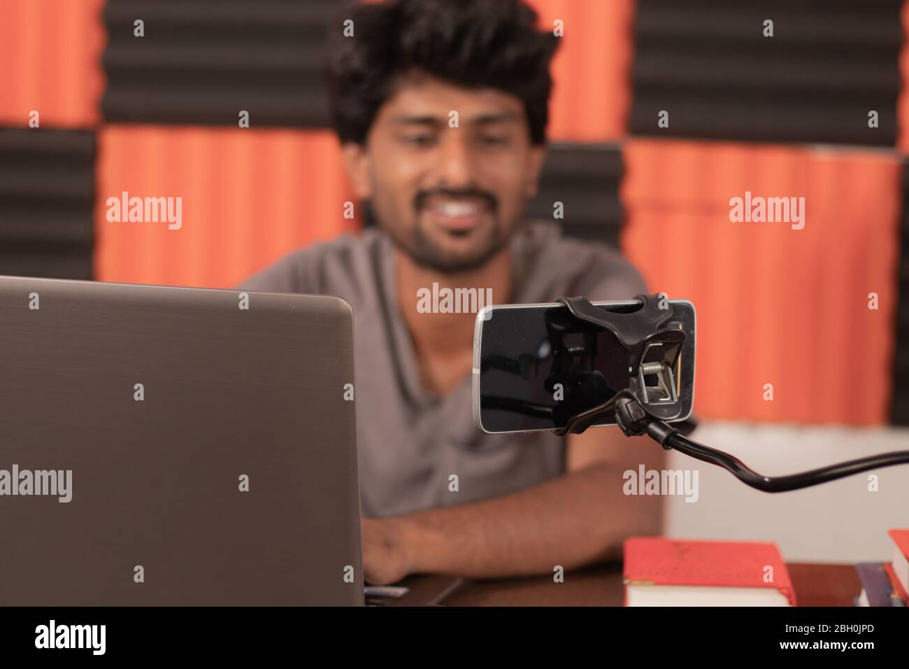 Junger Mann, der auf dem Handy beschäftigt im Video-Chat mit Freunden zu Hause während der covid-19 oder Coronavirus Pandemie - lächelnd Blogger mit Laptop-Filmen Stockfoto