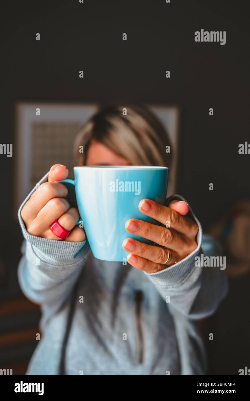 Frau, die in gemütlicher Atmosphäre eine blaue Tasse vor ihrem Gesicht hält. Stockfoto