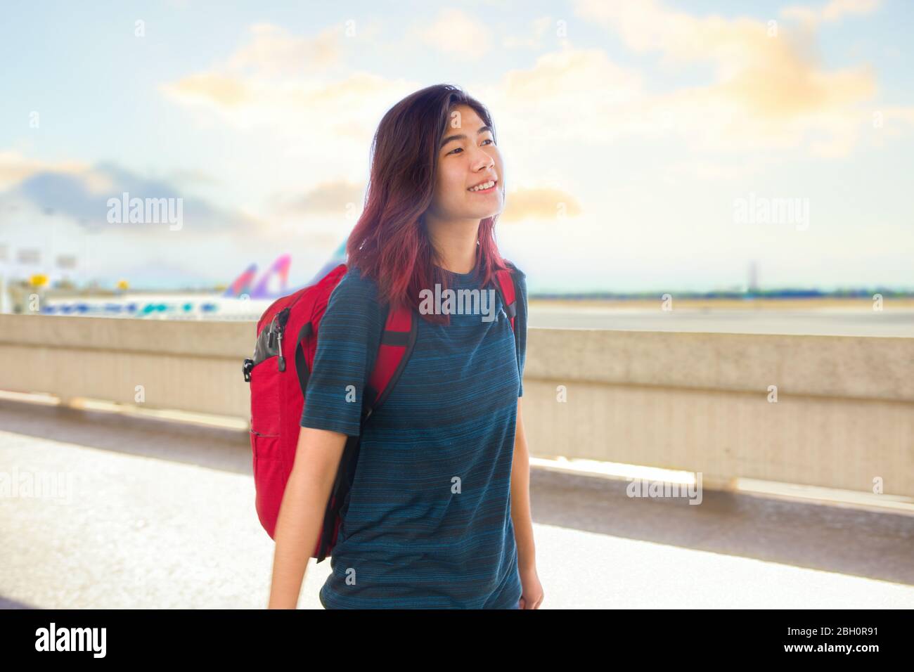 Lächelndes asiatisches Mädchen mit rotem Rucksack, das an hellen sonnigen Tagen am Flughafen an Reihen geparkter Flugzeuge vorbeigeht Stockfoto