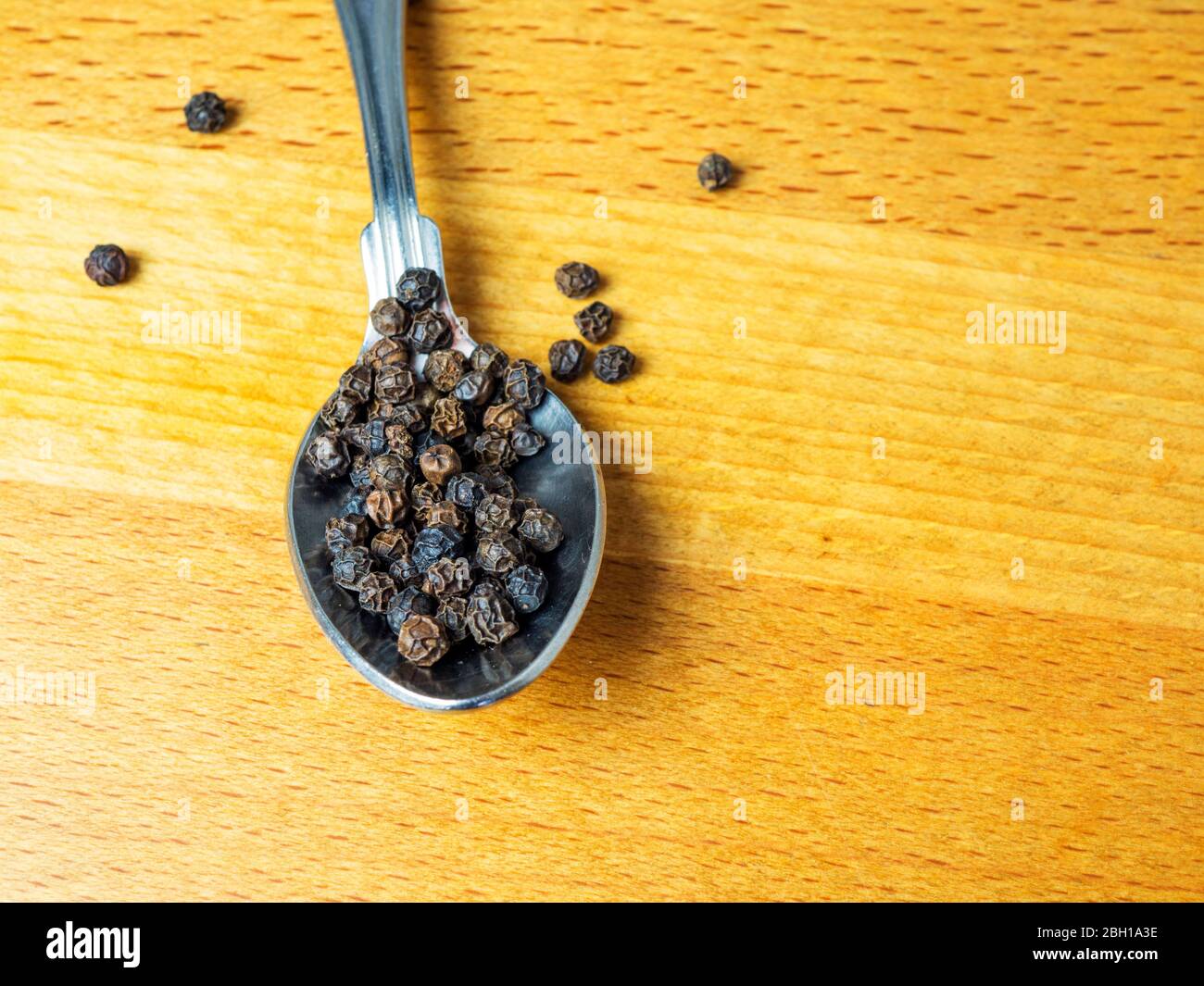 Ein Teelöffel schwarze Pfefferkörner auf einer Küchenarbeitspplatte, wobei einige auf die Holzoberfläche verschüttet werden Stockfoto