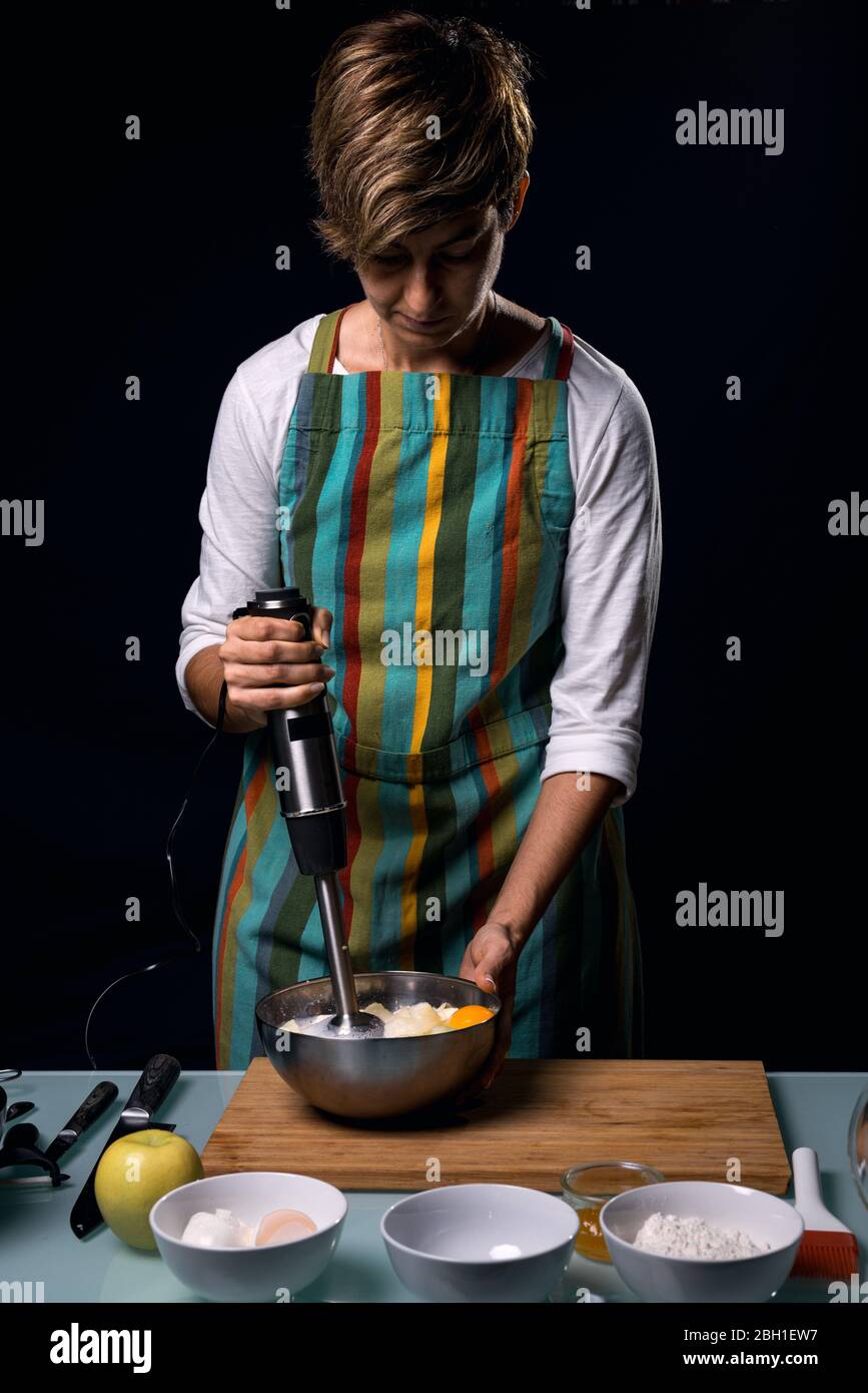 Arbeiten in der Küche. Schlagen Zutaten mit einem elektrischen Mixer. Kochen Apfelkuchen. Bunte Schürze. Kochtisch. Dunkelschwarzer Hintergrund. Vertikale Vie Stockfoto