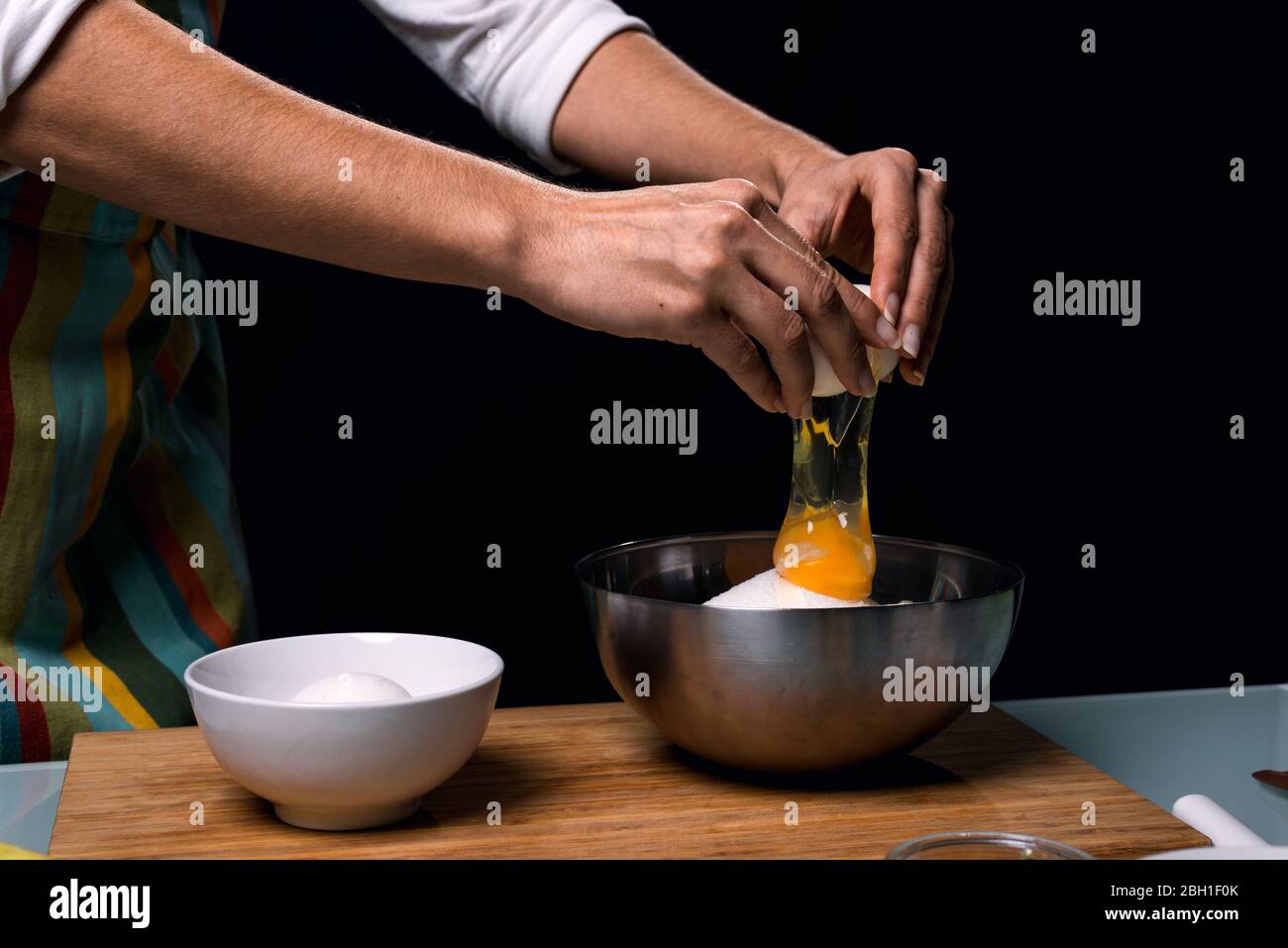 Arbeiten in der Küche. Ein Ei in einer Schüssel knacken. Kochen Apfelkuchen. Bunte Schürze. Kulinarische Zutaten. Kochtisch. Dunkelschwarzer Hintergrund. Stockfoto