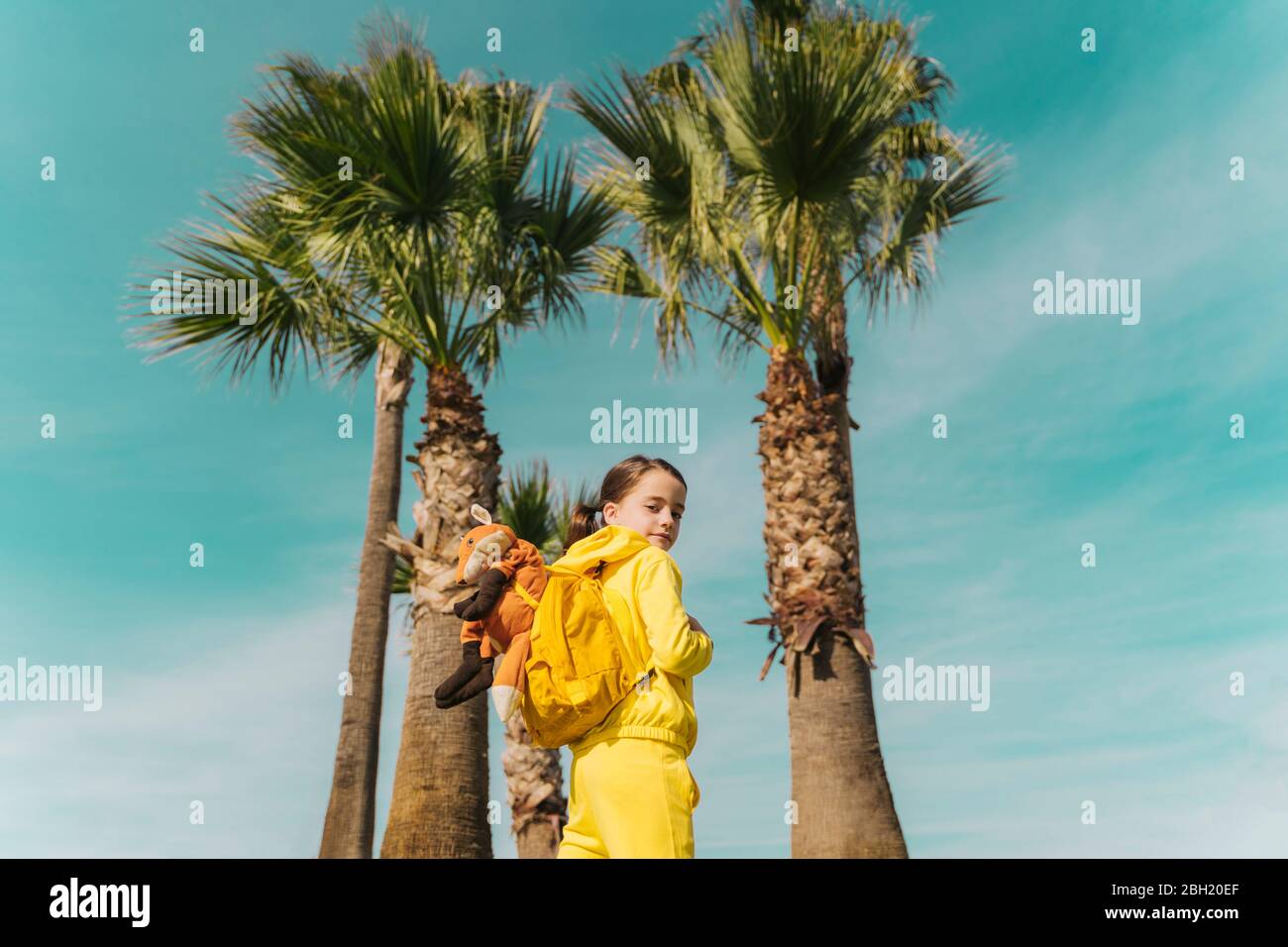 Portrait von kleinen Mädchen mit Rucksack und Plüsch vor Palmen Stockfoto